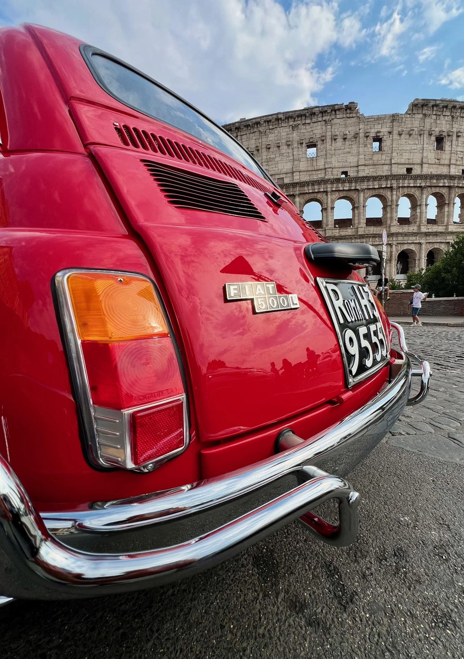 Auto Fiat 500L rossa parcheggiata di fronte al Colosseo a Roma, con persone che passeggiano sullo sfondo.