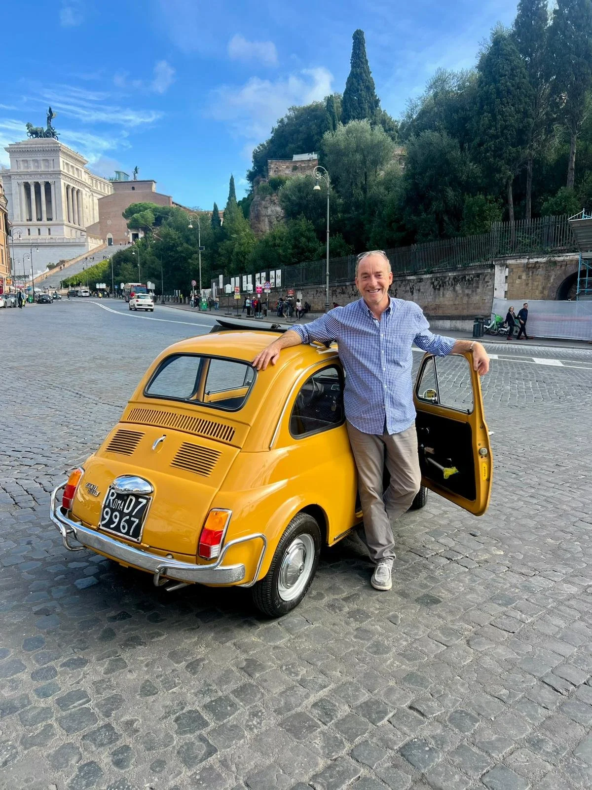 Uomo sorridente con camicia a quadretti grigia e beige pantaloni, che si appoggia sulla portiera di una piccola auto gialla d'epoca, parcheggiata su una piazza di ciottoli con sfondo di monumenti e alberi.