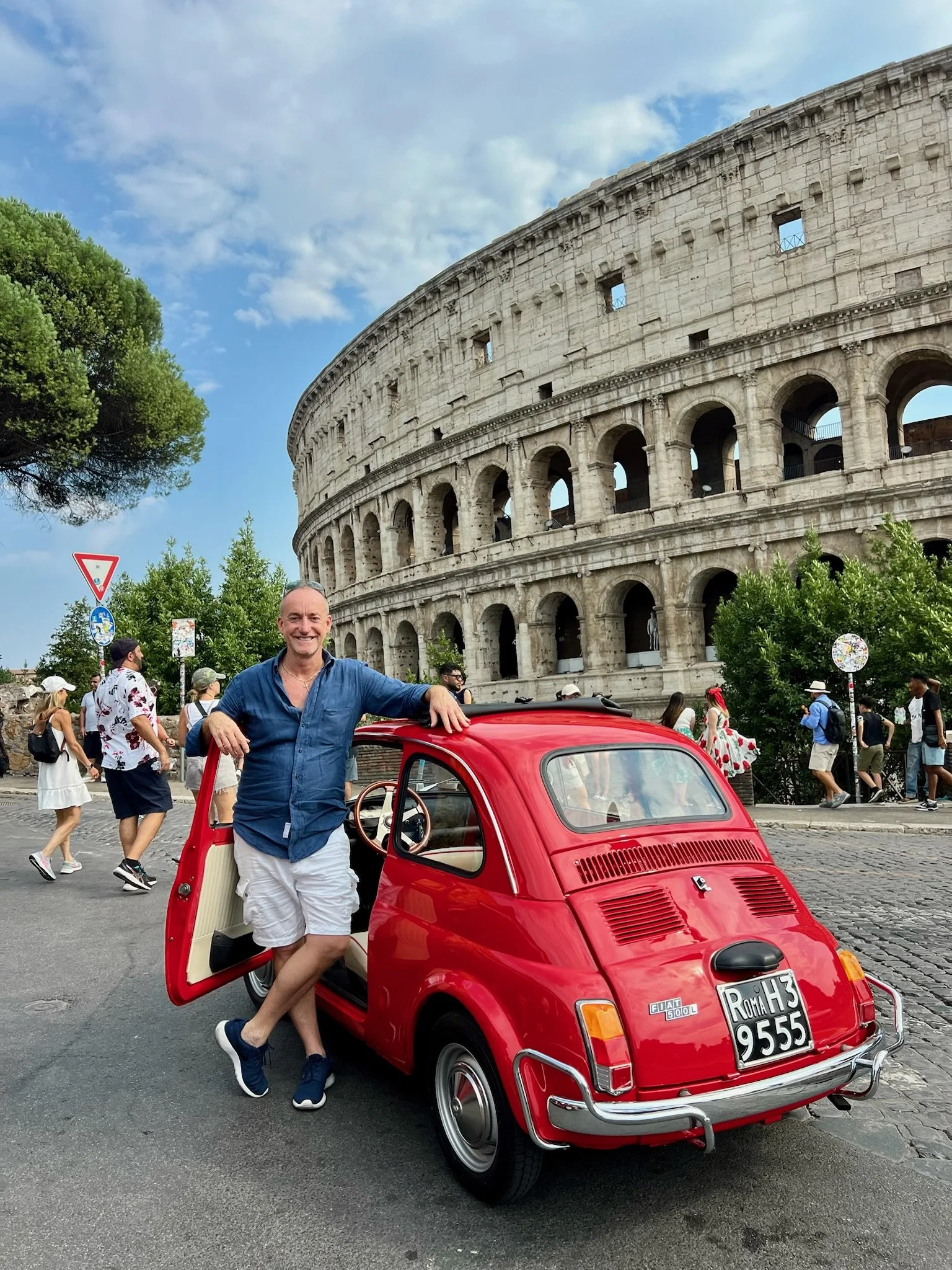 Uomo sorridente con camicia blu e pantaloncini bianchi è appoggiato a una piccola auto rossa Fiat 500 davanti al Colosseo a Roma. Ci sono persone che camminano dietro e il cielo è parzialmente nuvoloso.