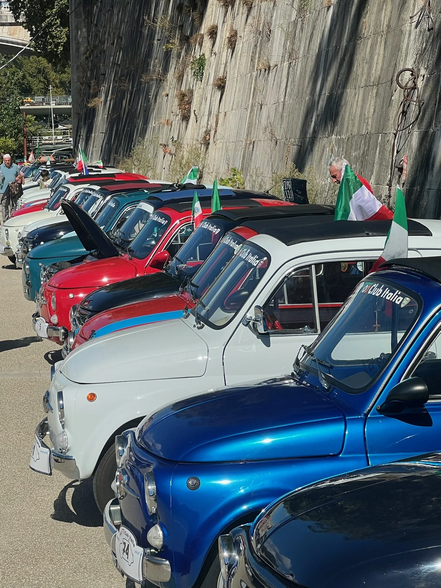 Una fila di auto d'epoca colorate, principalmente in bianco, rosso e blu, con alcuni partecipanti che sventolano bandiere italiane, sembra essere un raduno di auto storiche, vicino a un muro di roccia.