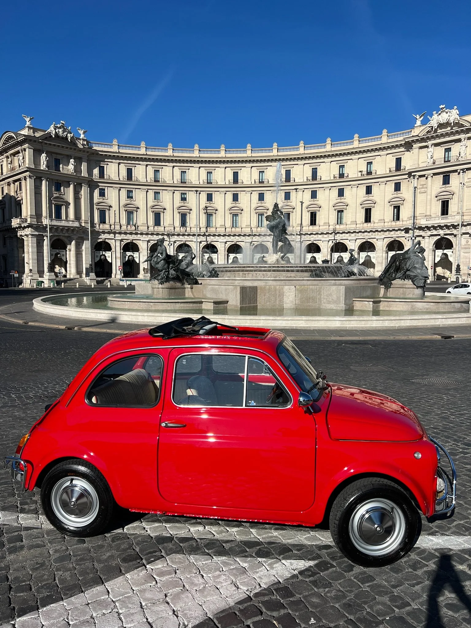 Una Fiat 500 rossa parcheggiata su una strada di ciottoli davanti a una fontana con sculture di cavalli e figure umane, con un edificio storico sullo sfondo e un cielo azzurro.