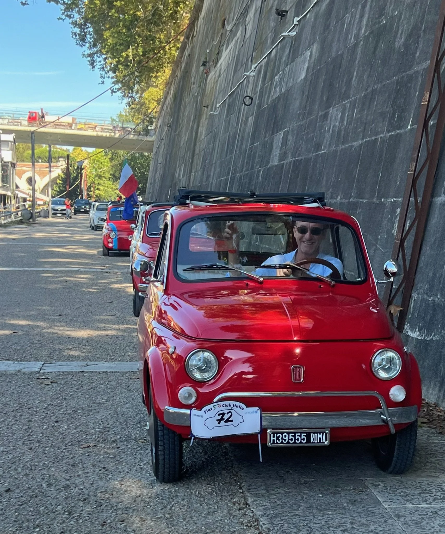 Una vettura rossa classica con una bandiera italiana e una targhetta con il numero 72, partecipante a un evento automobilistico a Roma, con un uomo sorridente che guida, indossa occhiali da sole e saluta.