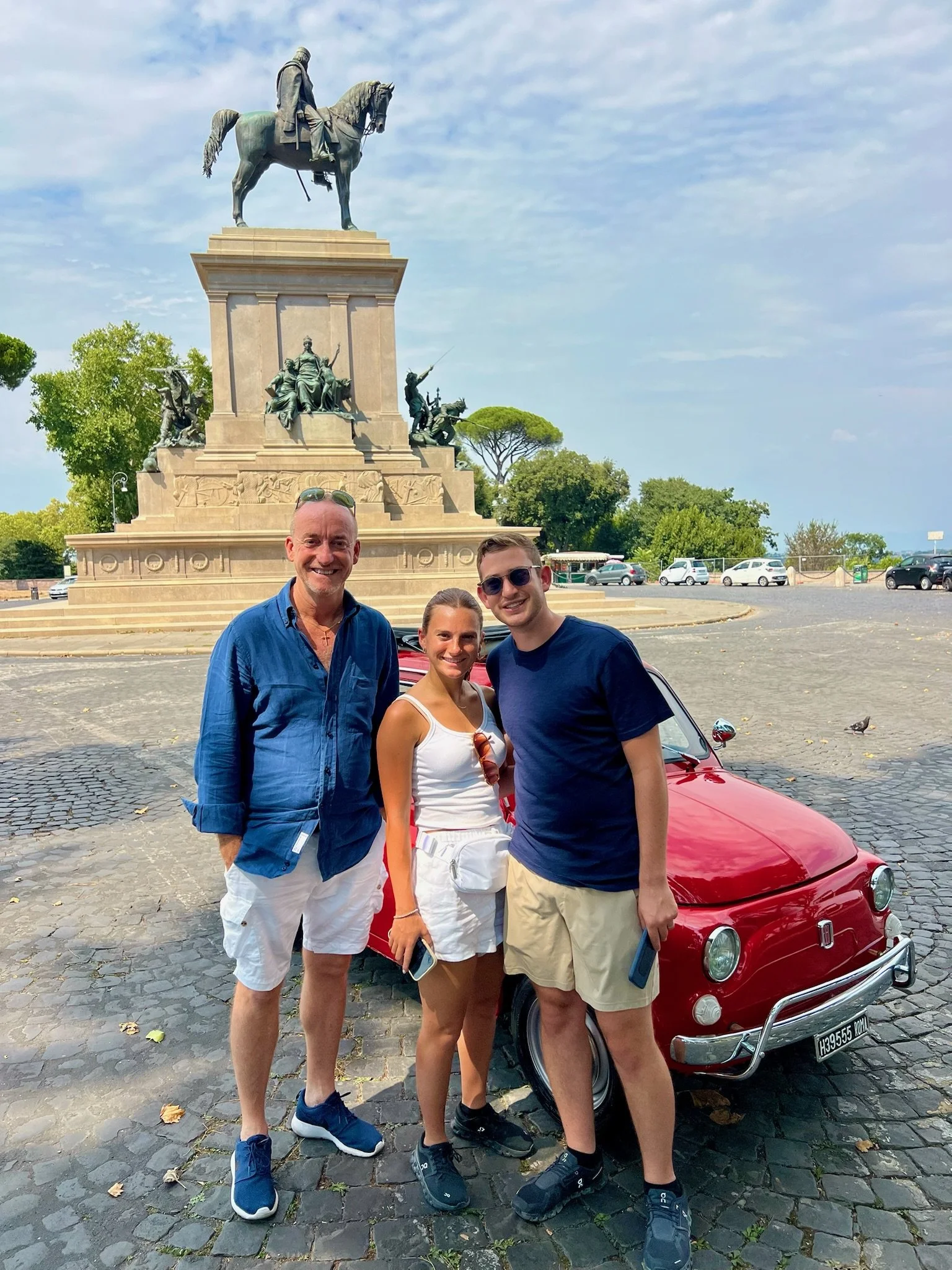Tre persone sorridenti davanti a una scultura e una macchina rossa vintage. La scultura è un monumento con figure e un cavaliere a cavallo sullo sfondo di un cielo nuvoloso e alberi verdi.