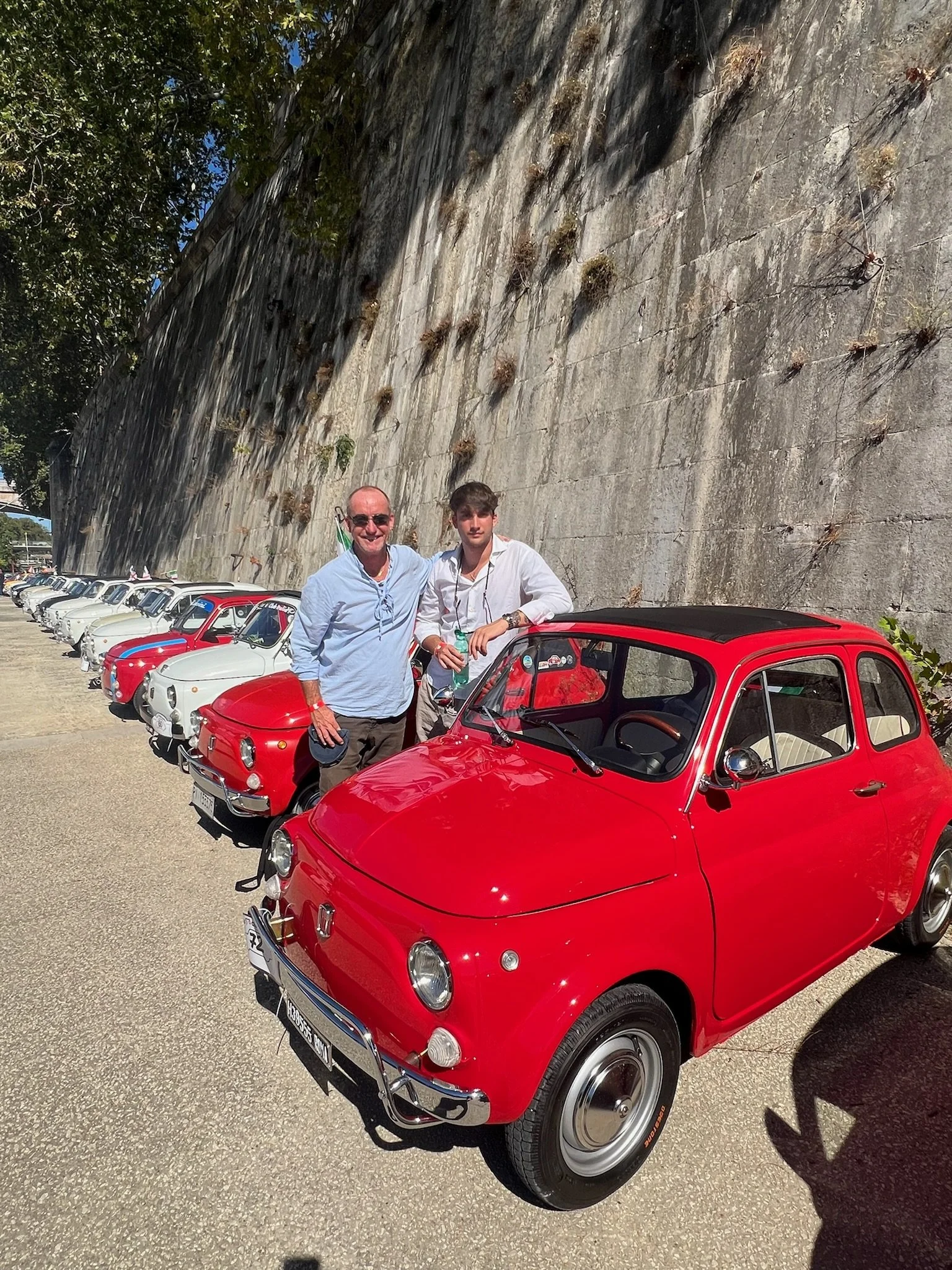 Due persone, un adulto e un adolescente, posano accanto a un'auto rossa in una fila di auto storiche bianche e rosse, con un muro in pietra sullo sfondo in una giornata soleggiata.