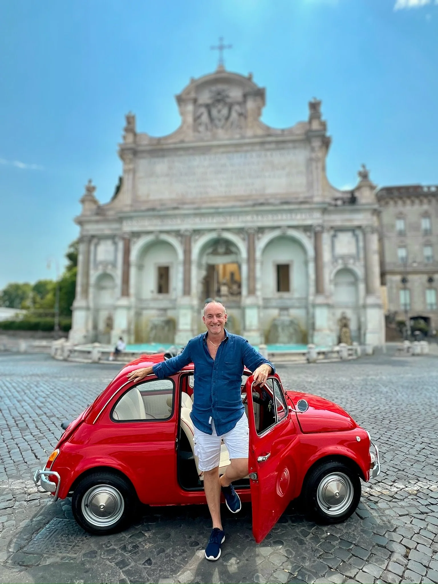 Uomo sorridente con camicia blu e pantaloncini bianchi in piedi accanto a una piccola auto rossa in una piazza con un edificio storico in background.