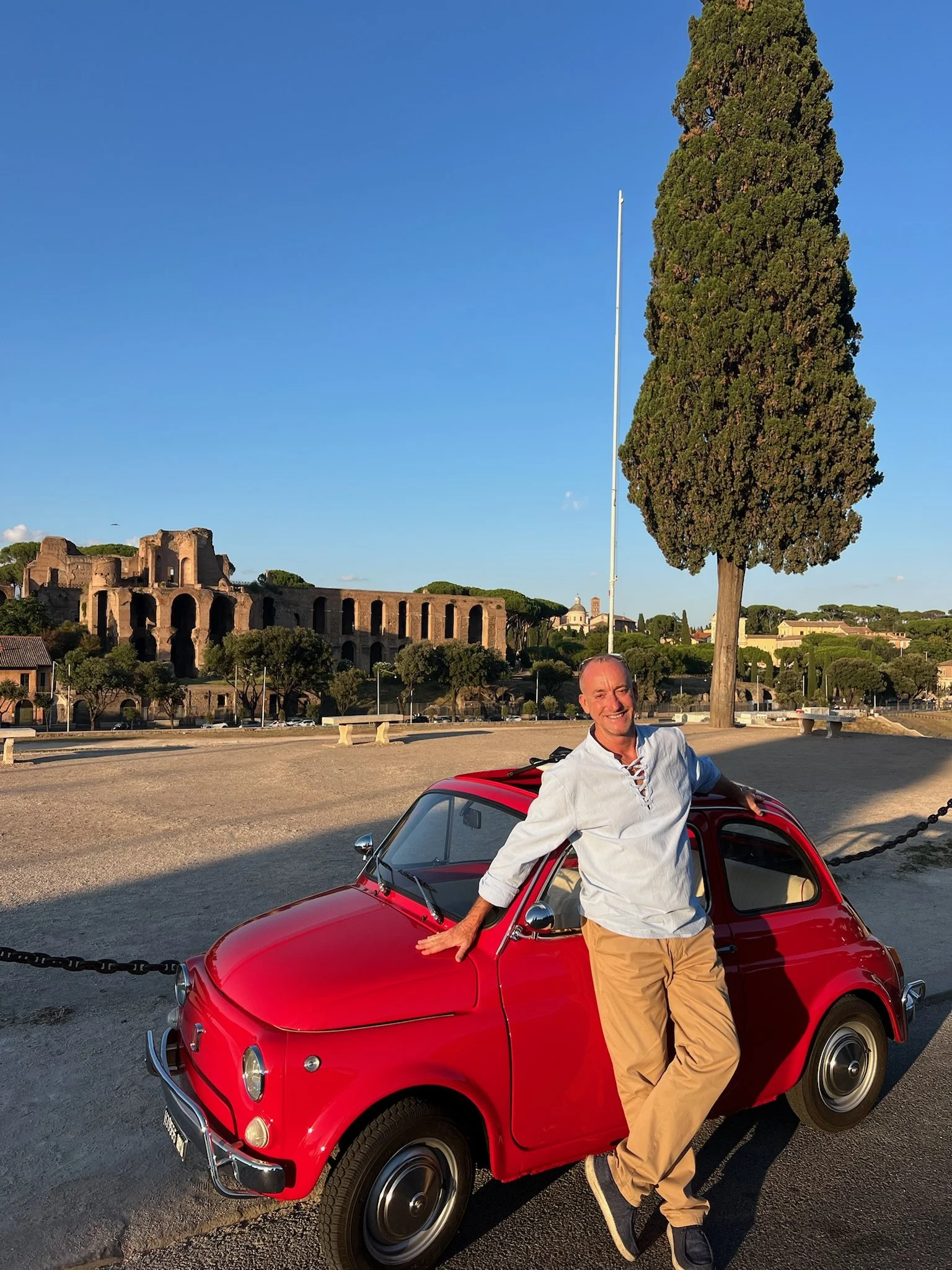 Uomo sorridente che si appoggia a una piccola auto rossa in un'area all'aperto con un grande albero e rovine antiche sullo sfondo durante il tramonto.