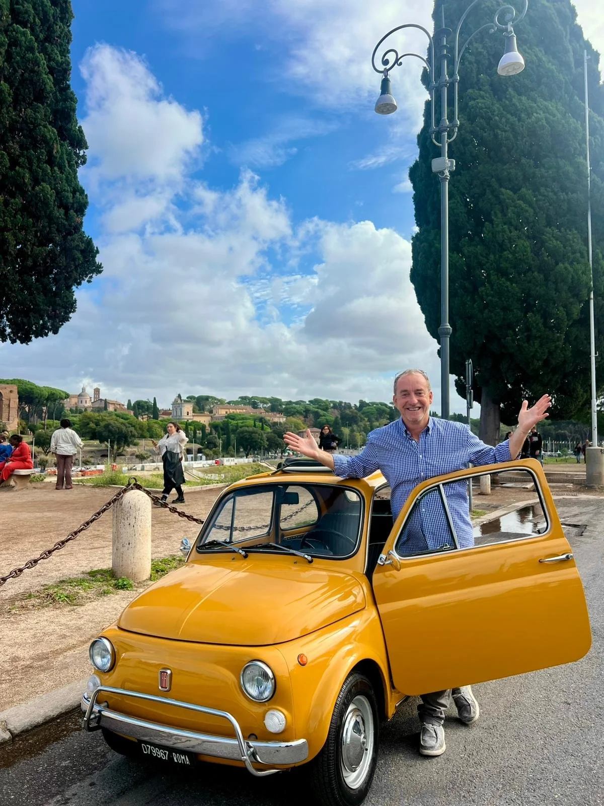 Un uomo felice in piedi accanto a una piccola auto gialla, con la porta aperta, in una zona aperta con alberi e persone sullo sfondo, in una giornata soleggiata con nuvole nel cielo.