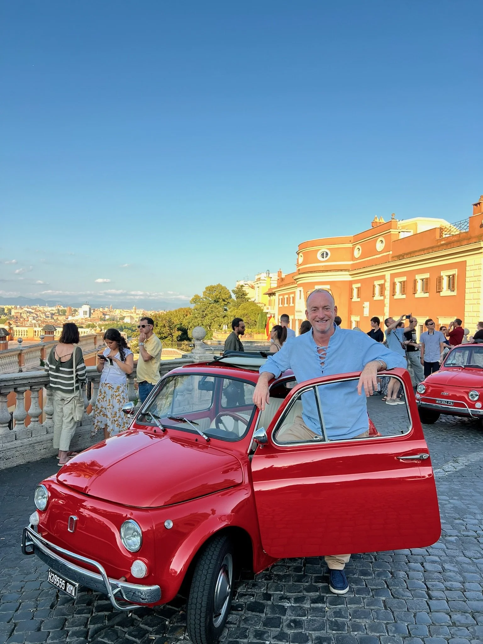 Un uomo sorridente si appoggia su una Fiat 500 rossa con lo sfondo di un gruppo di persone e un edificio storico in un'area urbana.