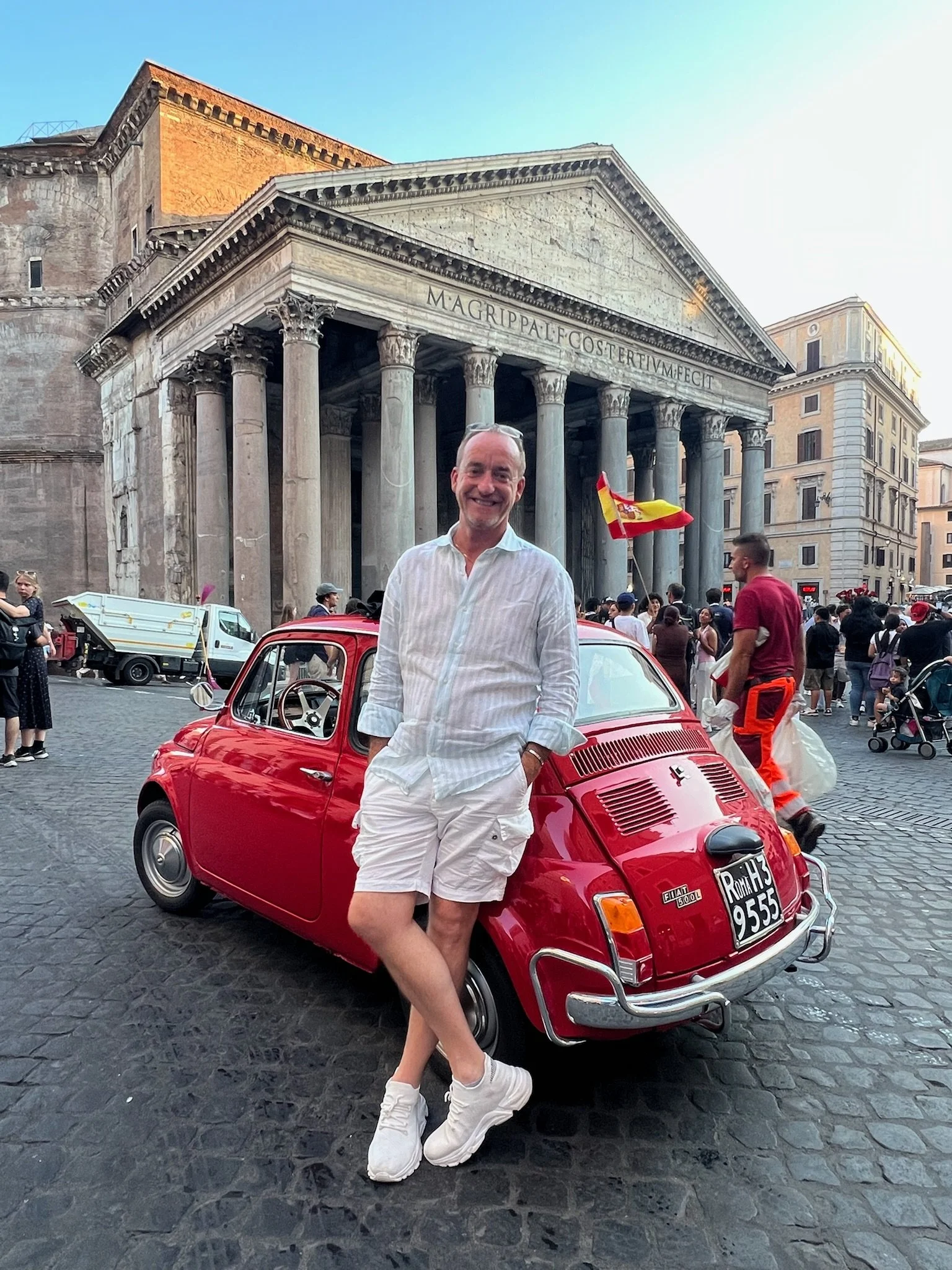 Uomo sorridente in abbigliamento bianco, con cane sul ginocchio, in piedi accanto a una Fiat 500 rossa, con il Colosseo sullo sfondo a Roma.
