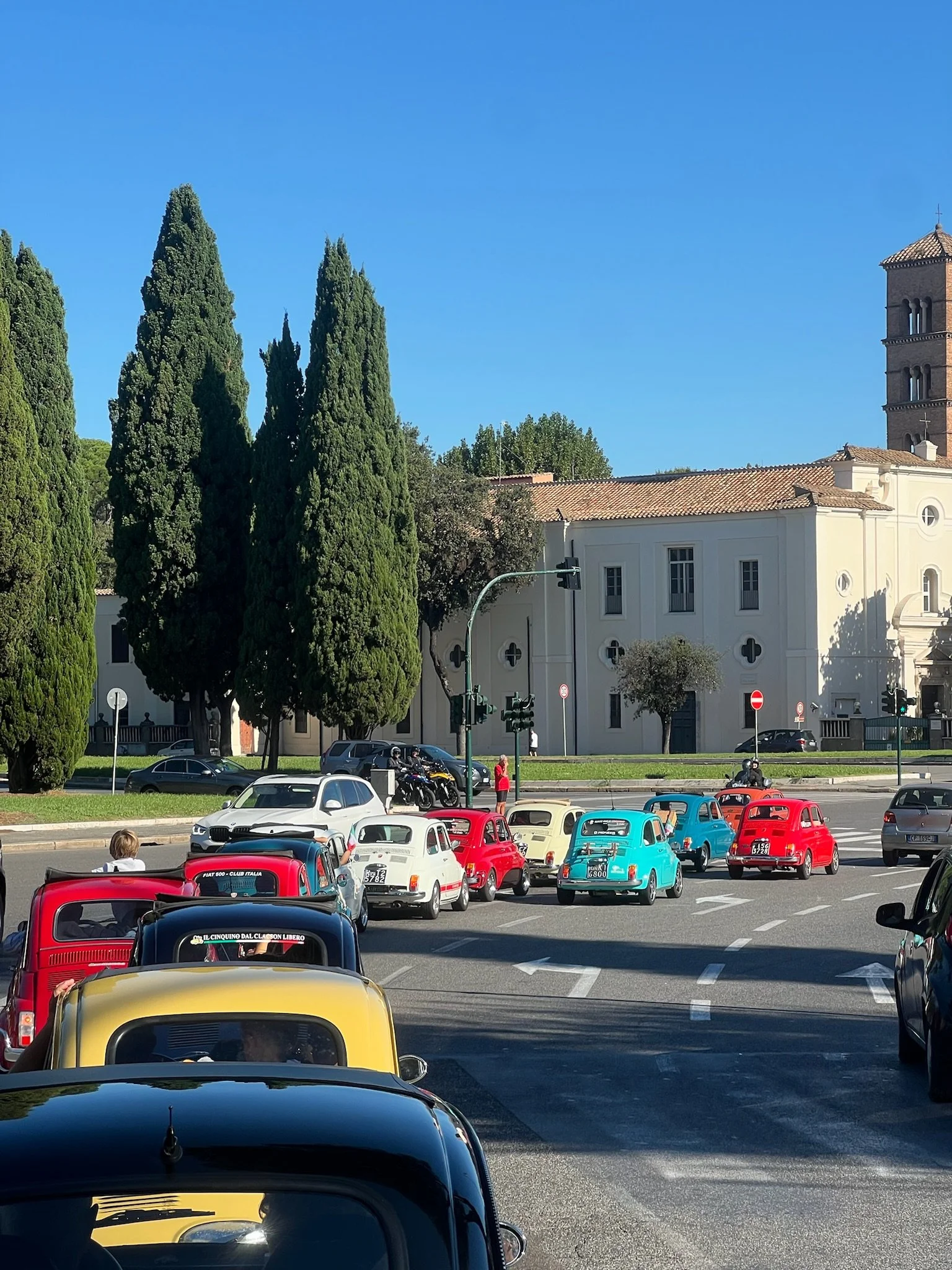 Una fila di piccole auto vintage di vari colori parcheggiate lungo la strada davanti a una chiesa bianca con torri e grandi alberi, sotto un cielo azzurro.