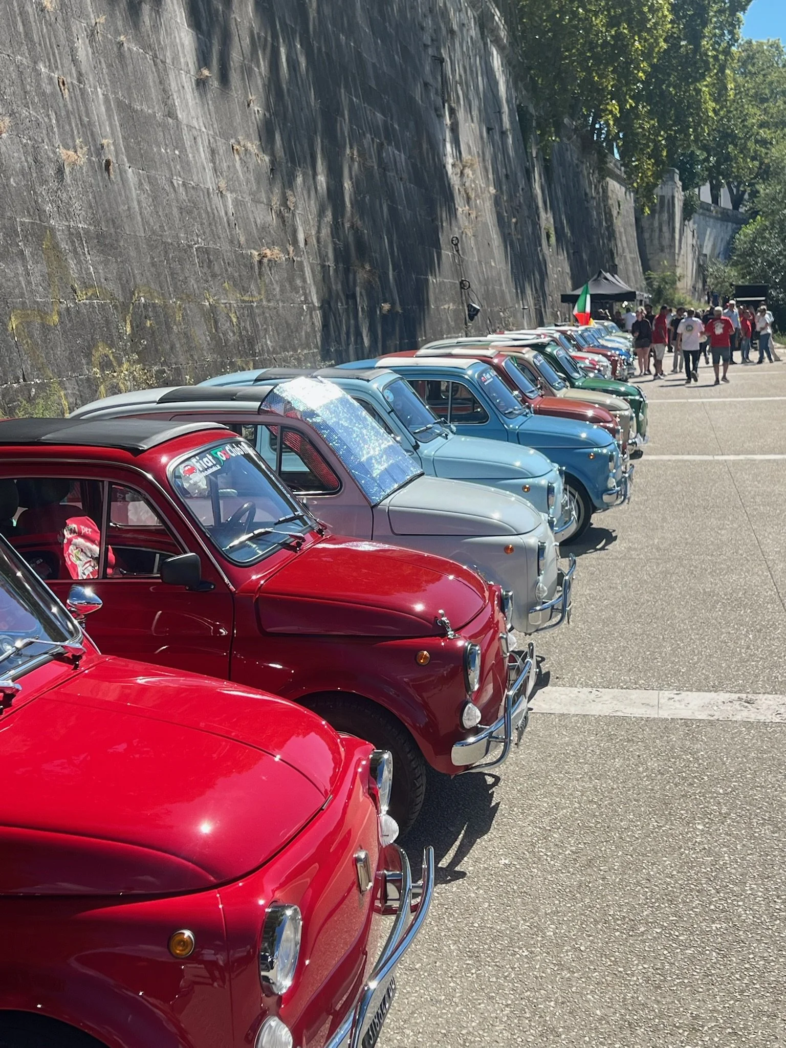 Fila di auto d'epoca colorate parcheggiate lungo un muro di pietra con alcune persone che passeggiano sullo sfondo