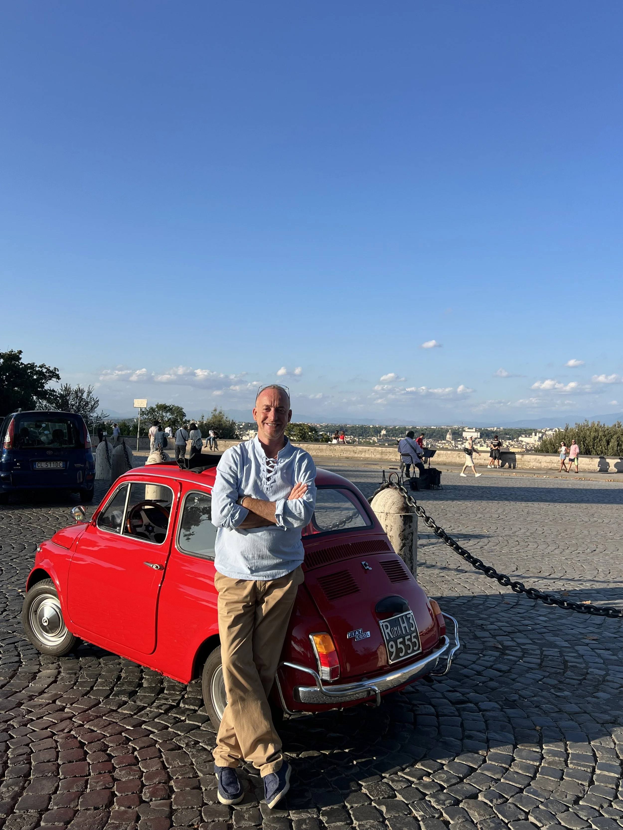 Uomo sorridente che si appoggia a una piccola auto rossa in un'area pedonale con molte persone e un cielo sereno sullo sfondo.