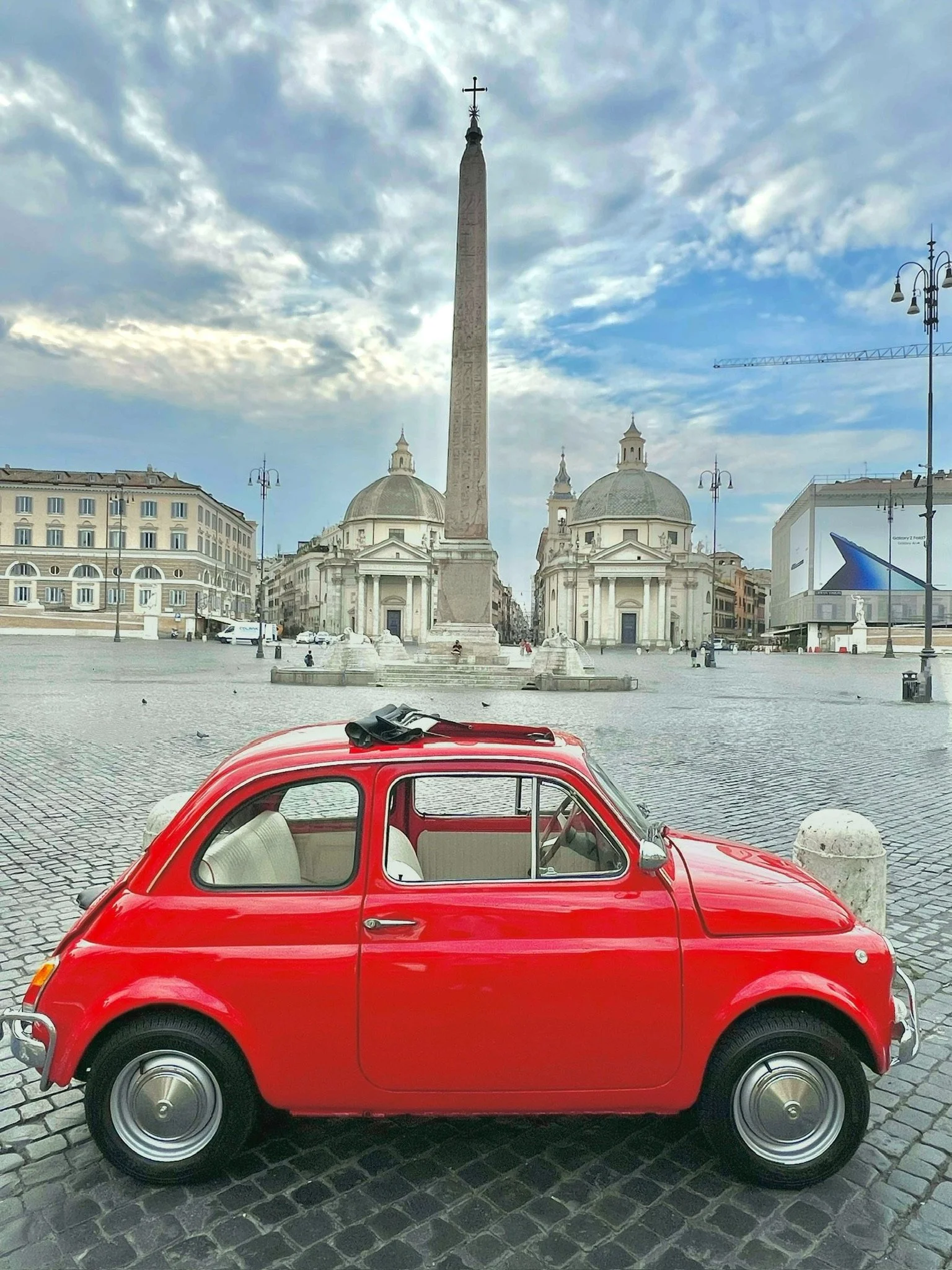 Auto rossa vintage parcheggiata in una piazza di Roma con un obelisco e chiese barocche sullo sfondo, cielo nuvoloso.