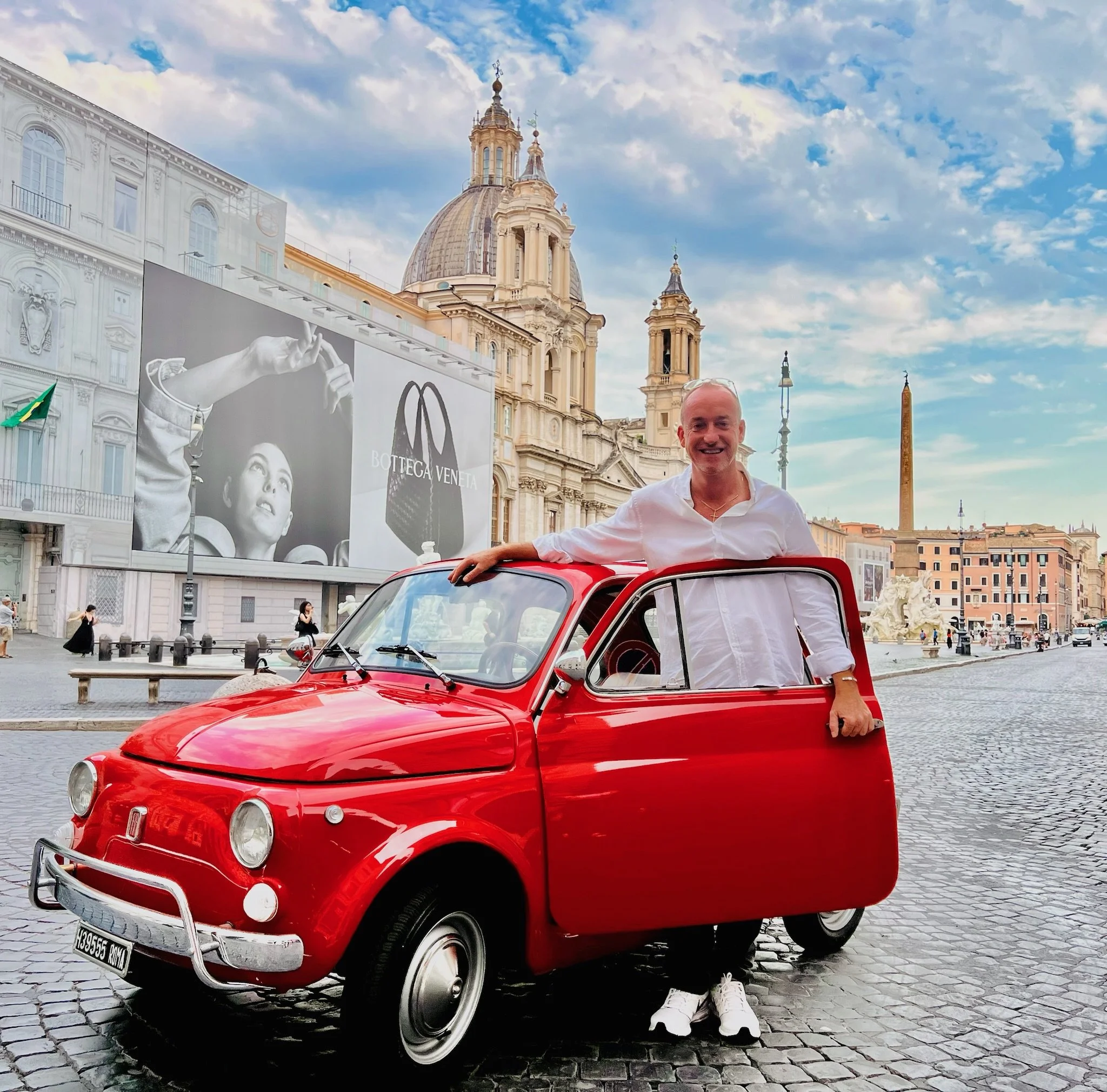 Uomo sorridente con camicia bianca si appoggia a una piccola Fiat rossa, in una piazza di Roma con edifici storici e il Colosseo sullo sfondo.