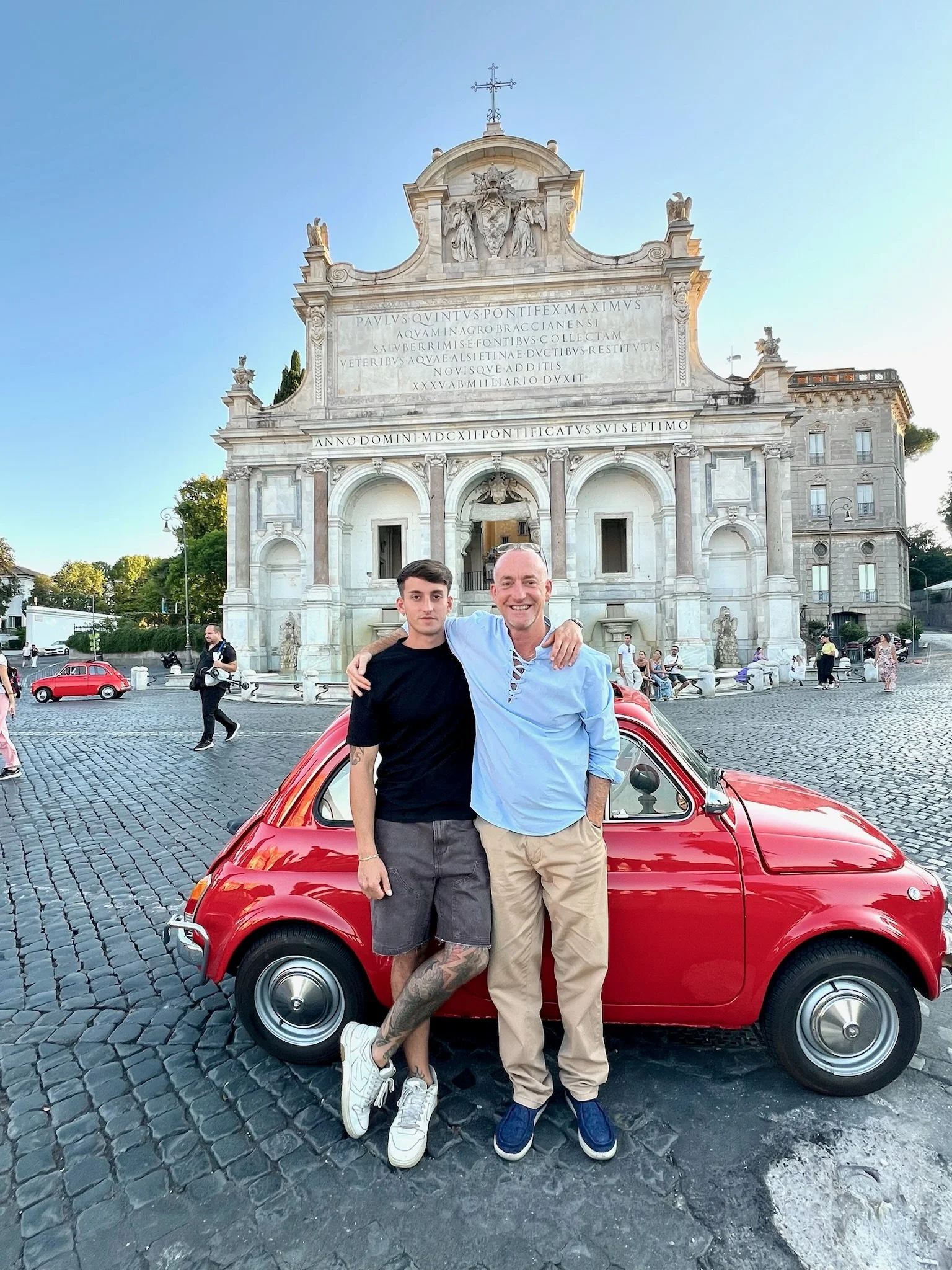 Due uomini sorridenti che si abbracciano davanti a una piccola auto rossa, con un edificio storico sullo sfondo, in piazza a Roma.
