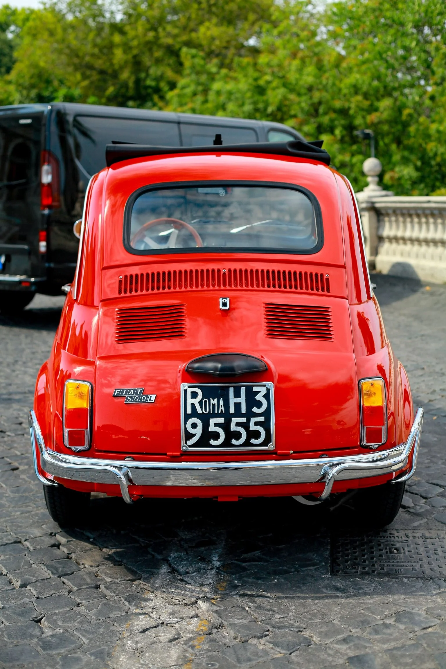 Fiat 500L rossa vista da dietro, parcheggiata su strada in pietra, con sfondo di alberi verdi e parte di un ponte in pietra.