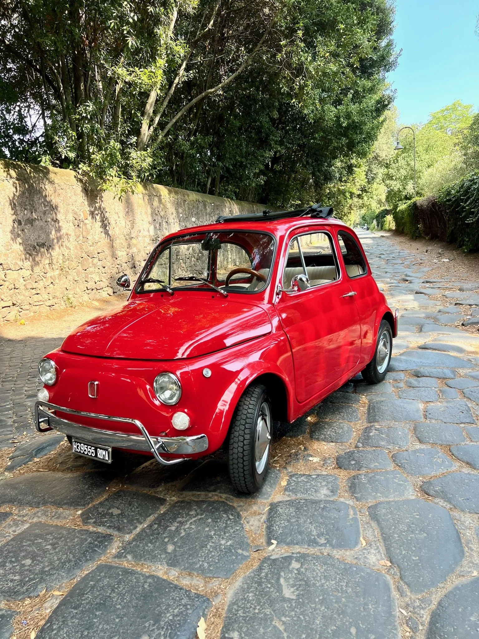 Auto d'epoca rossa parcheggiata su una strada di pietra con alberi e muro in pietra ai lati.