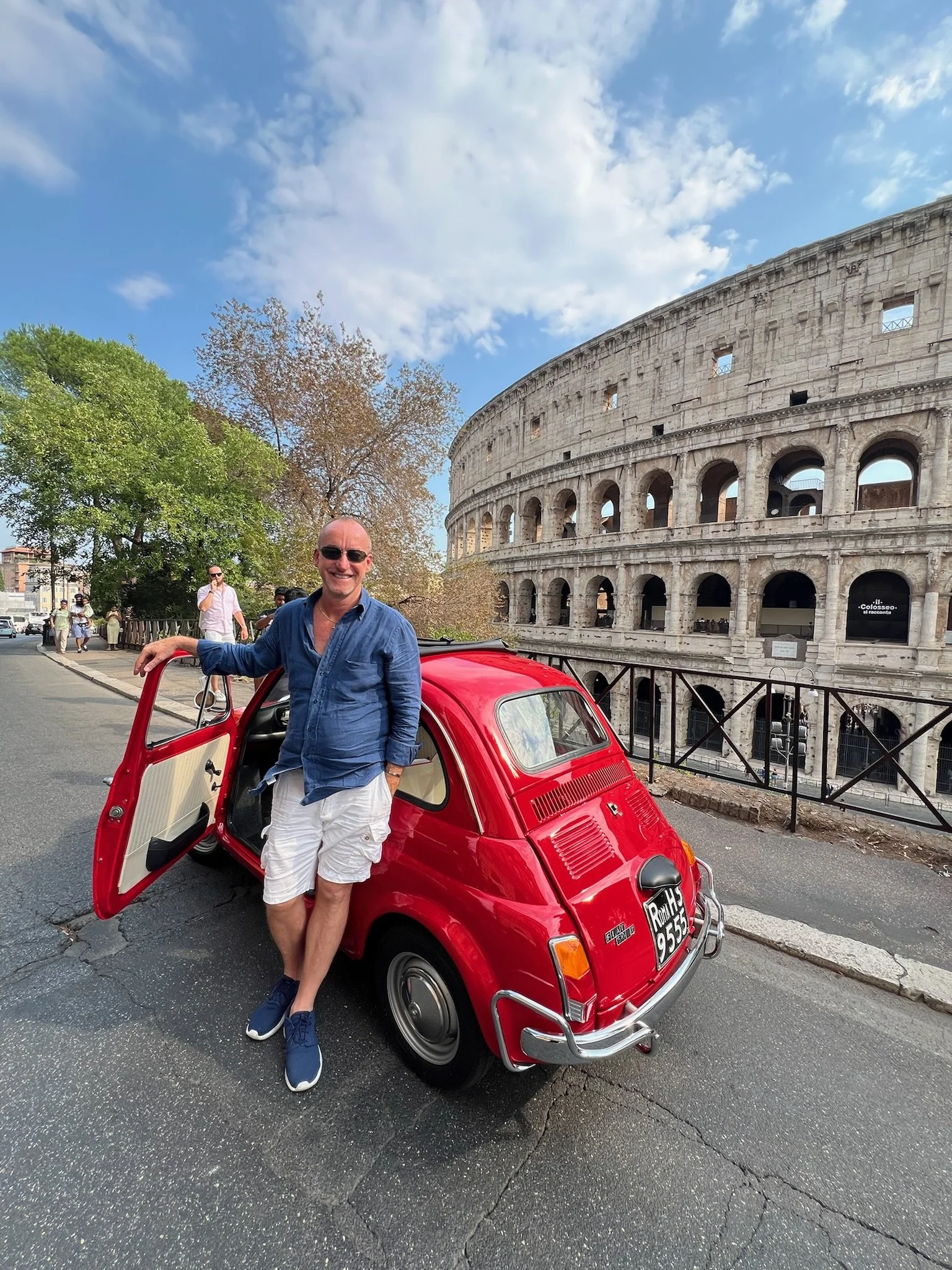 Uomo sorridente in abbigliamento casual posizionato accanto a una Fiat 500 rossa, con il Colosseo sullo sfondo a Roma, cielo nuvoloso e alcune persone in lontananza.