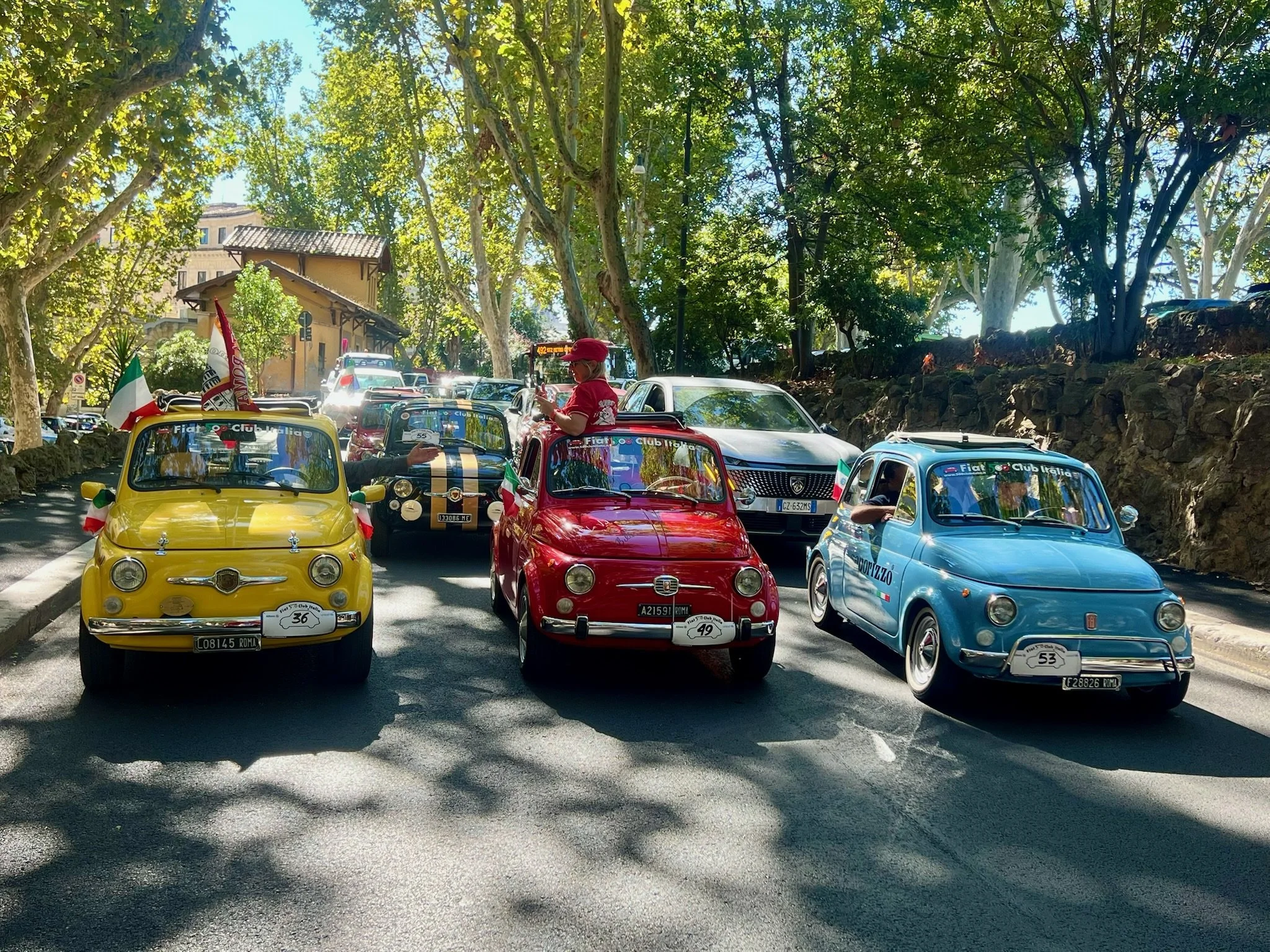 Tre auto d'epoca colorate, gialla, rossa e blu, parcheggiate sulla strada tra alberi e stabilimenti italiani, con persone vestite in stile vintage e bandiere italiane.