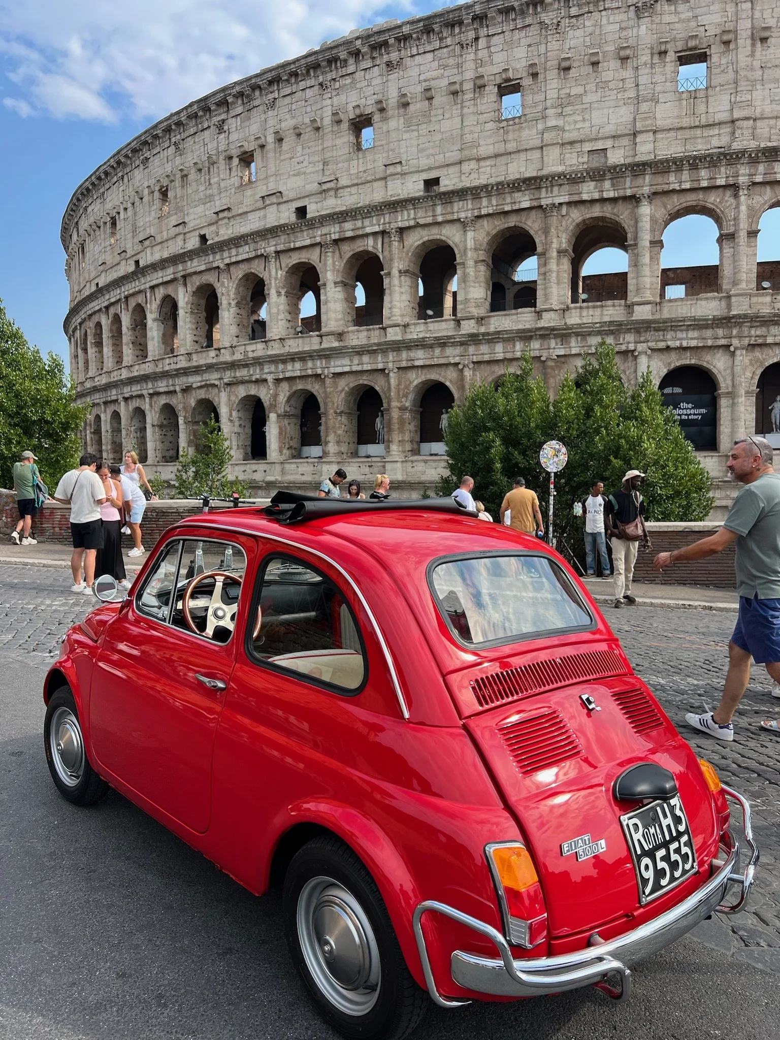 Una vecchia auto rossa Fiat 500 davanti al Colosseo a Roma, con turisti e visitatori in background e un cielo parzialmente nuvoloso.