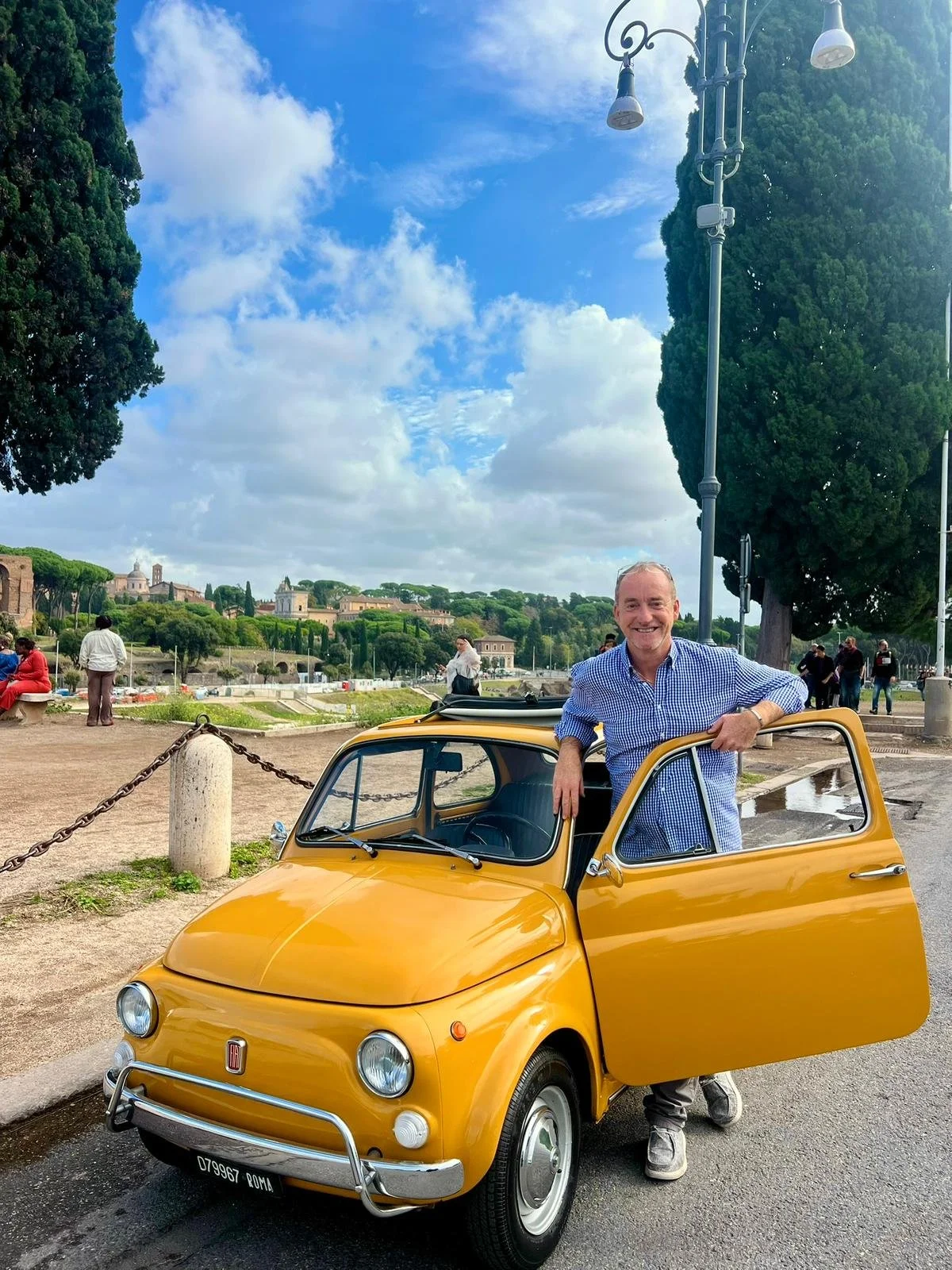 Uomo sorridente con camicia a quadri blu e bianca si appoggia a una piccola auto gialla vintage, sopra strada asfaltata con alcuni alberi e persone sullo sfondo, cielo con nuvole e alberi alti.