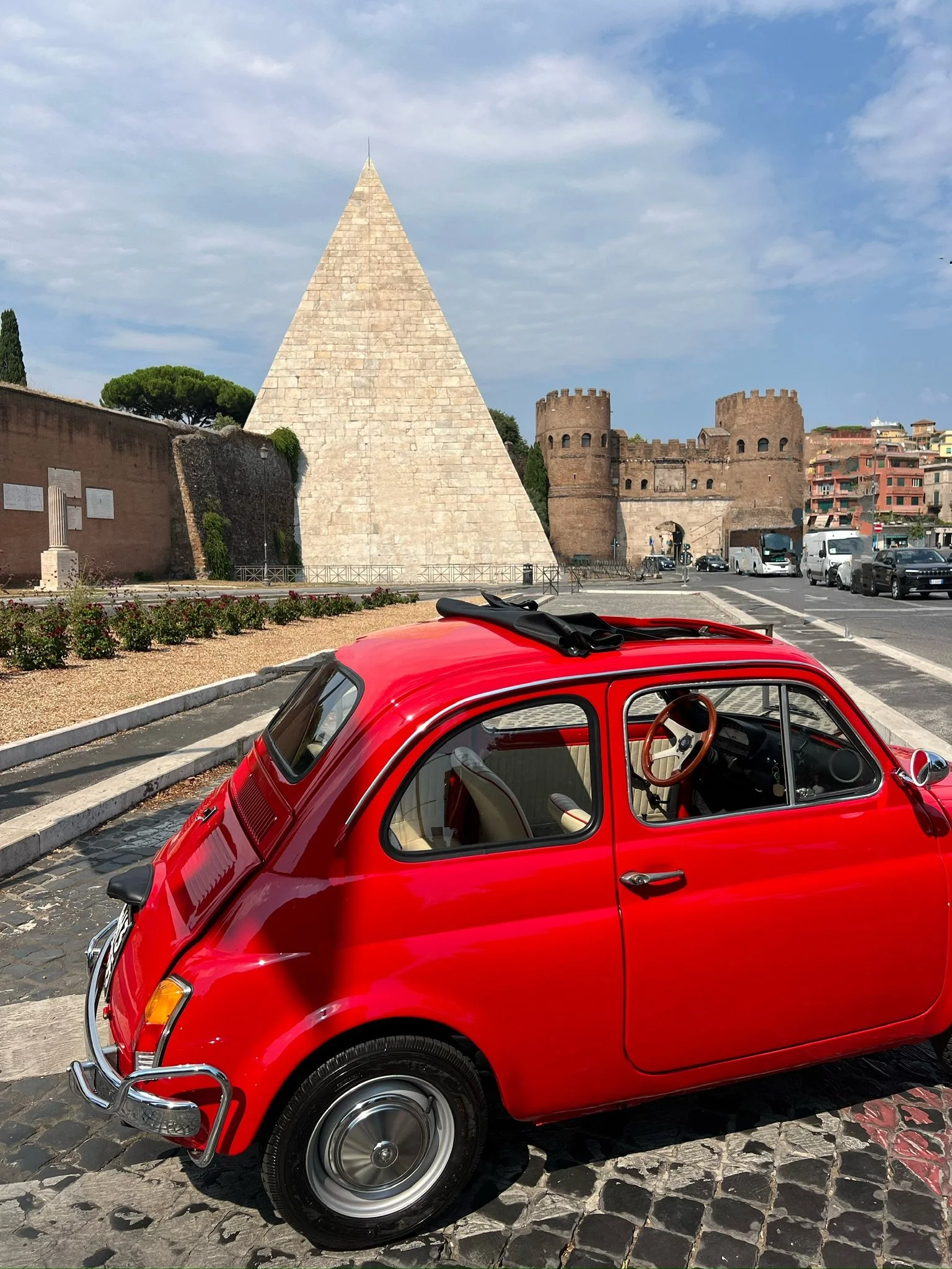Auto rosso vintage parcheggiata su strada con monumenti storici e edifici in background.