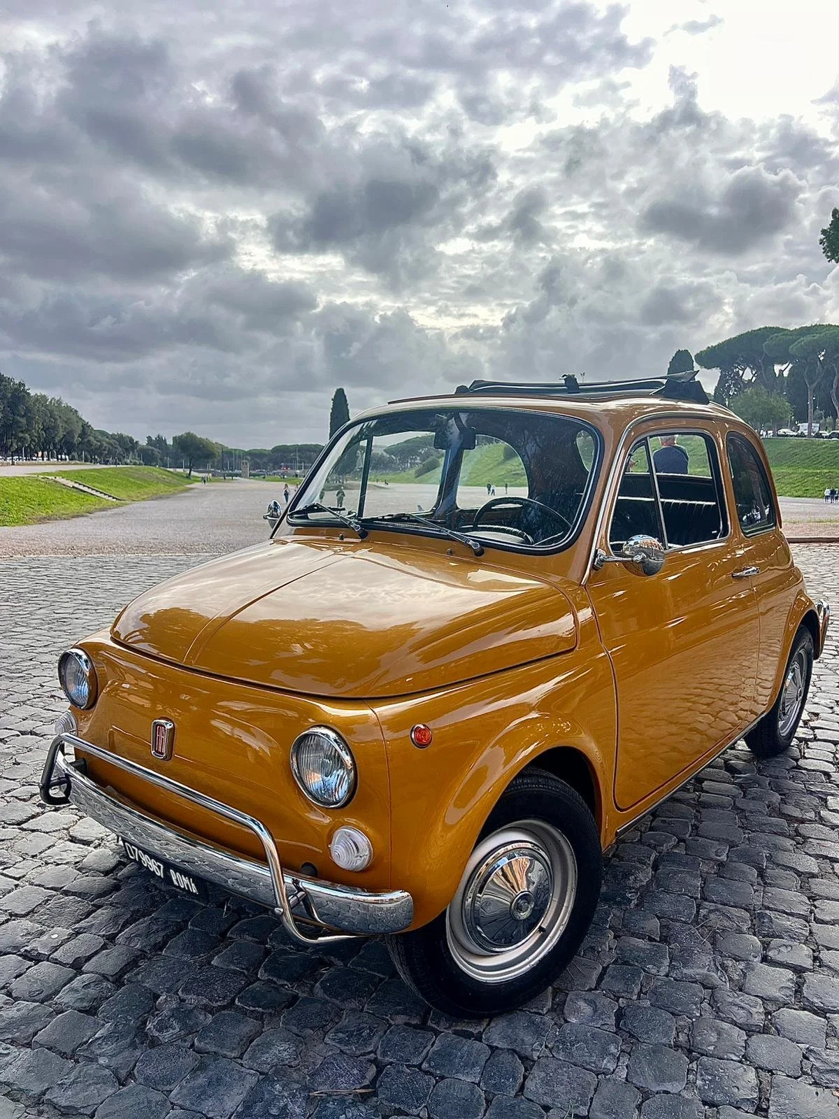 Auto d'epoca Fiat 500 di colore giallo, parcheggiata su una strada di ciottoli, con cielo nuvoloso sullo sfondo.