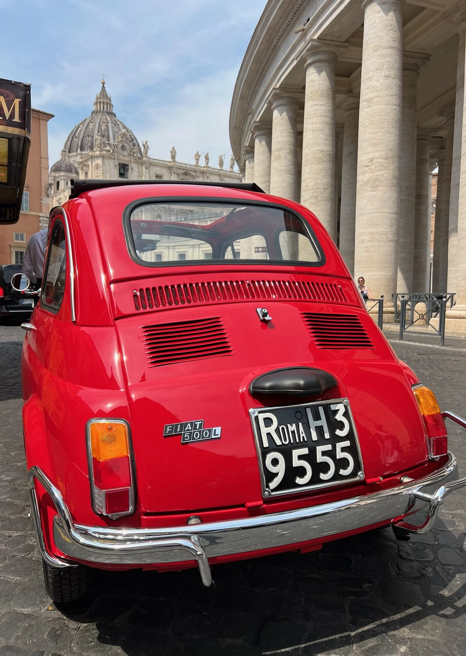 Auto vintage rossa Fiat 500L con targa Roma, parcheggiata davanti a un edificio storico con colonne e cupola in stile barocco.