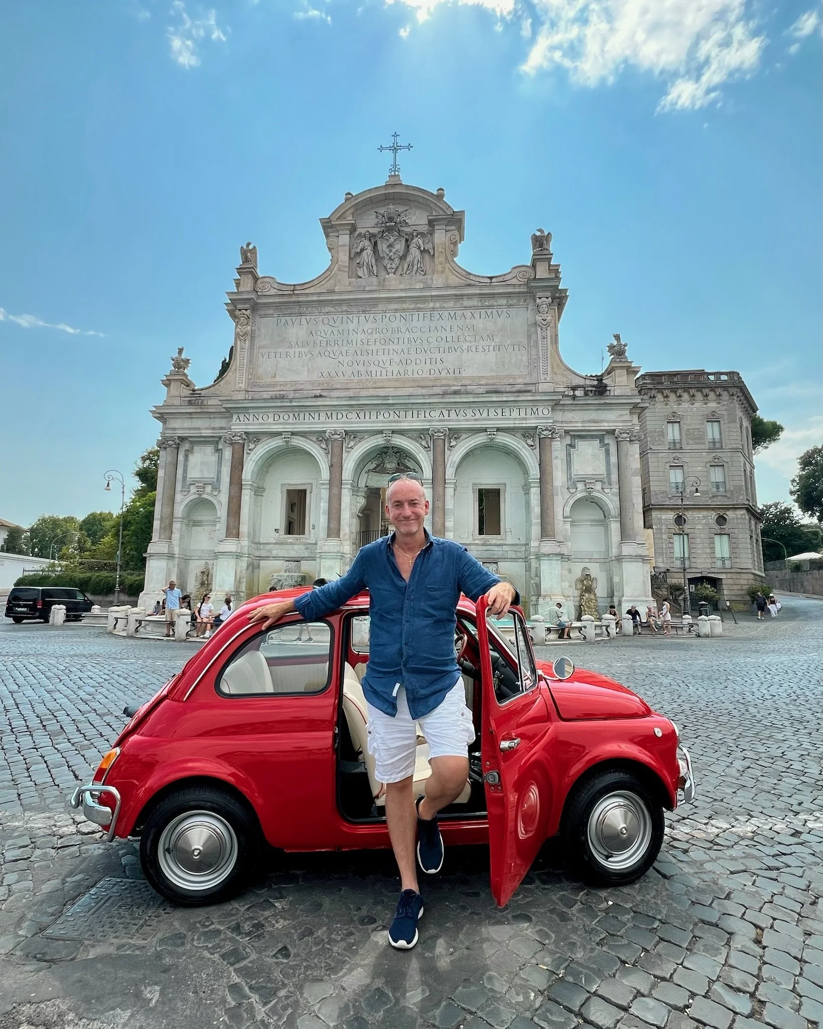 Uomo sorridente con camicia blu e pantaloncini bianchi in piedi accanto a una piccola auto rossa, con lo sfondo di un edificio storico in stile barocco in piazza, cielo blu e poche nuvole.