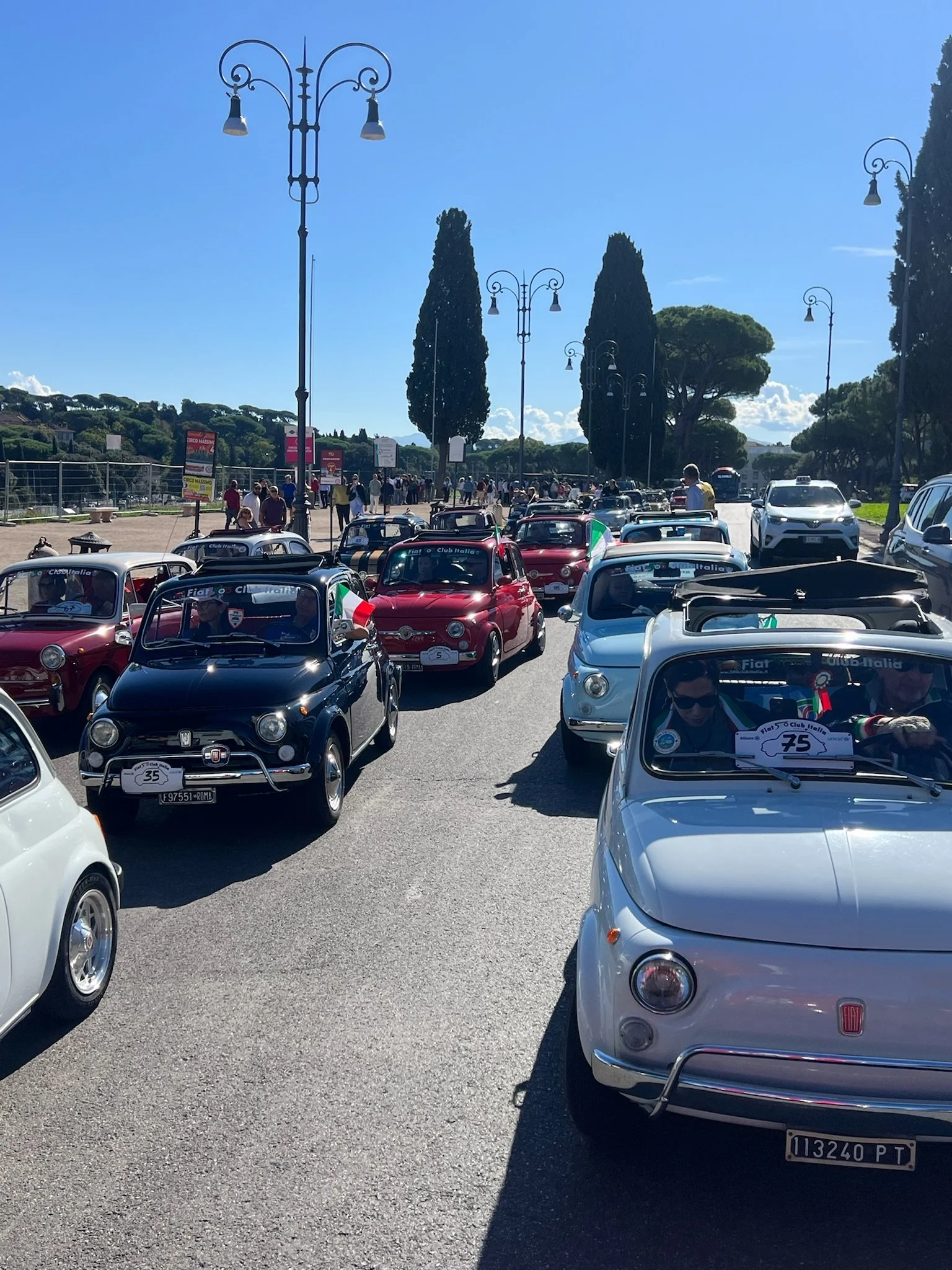 Ritrovo di molte automobili d'epoca Fiat, alcune con bandiere e adesivi in occasione di un raduno, con persone che camminano sul lato destro e luci stradali alte lungo la strada, sotto un cielo blu con poche nuvole.