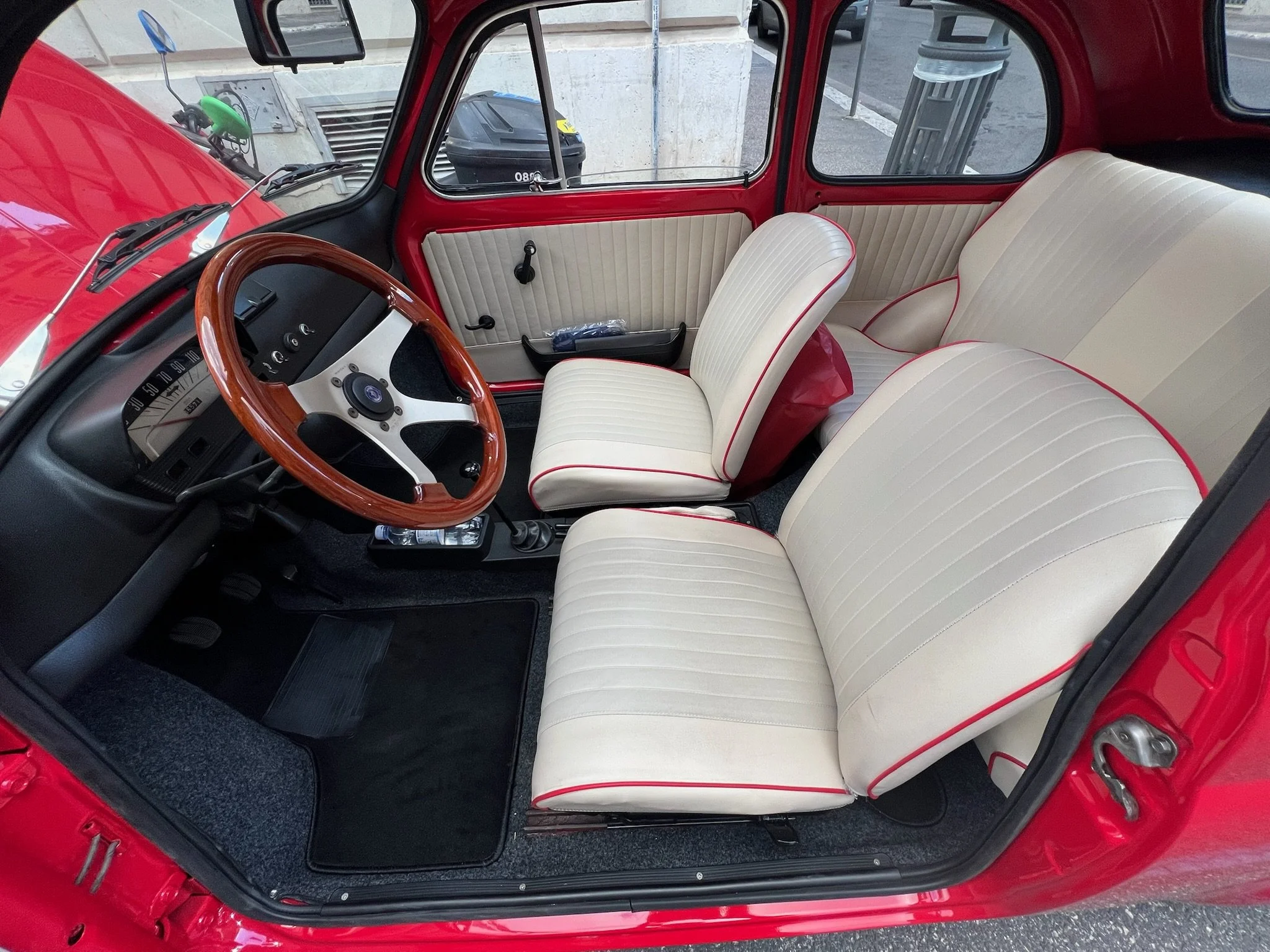 Interno di un'auto d'epoca rossa, con sedili beige con cuciture rosse, volante in legno e cruscotto nero.