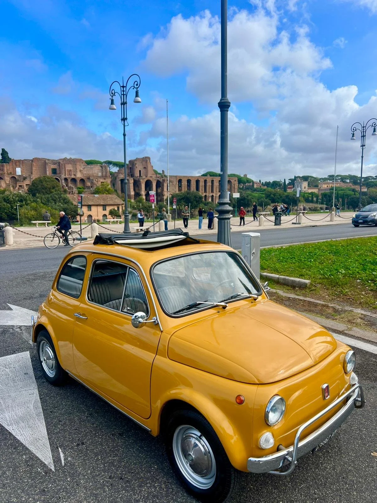 Auto d'epoca gialla parcheggiata su strada in Roma, con rovine antiche e persone in background, cielo nuvoloso.
