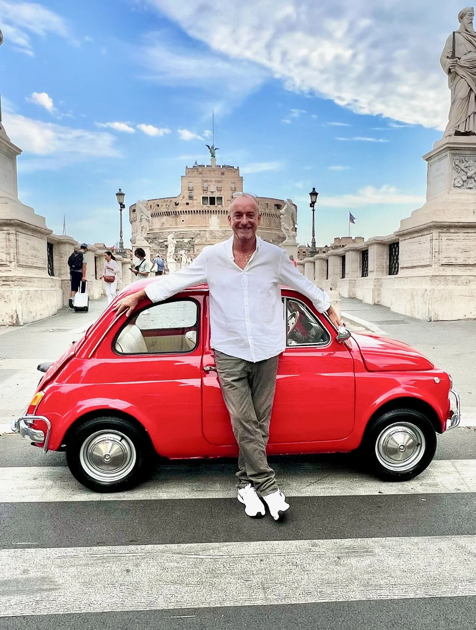 Uomo sorridente che si appoggia a una piccola auto rossa in un piazzale con monumenti storici sullo sfondo, probabilmente in Italia.