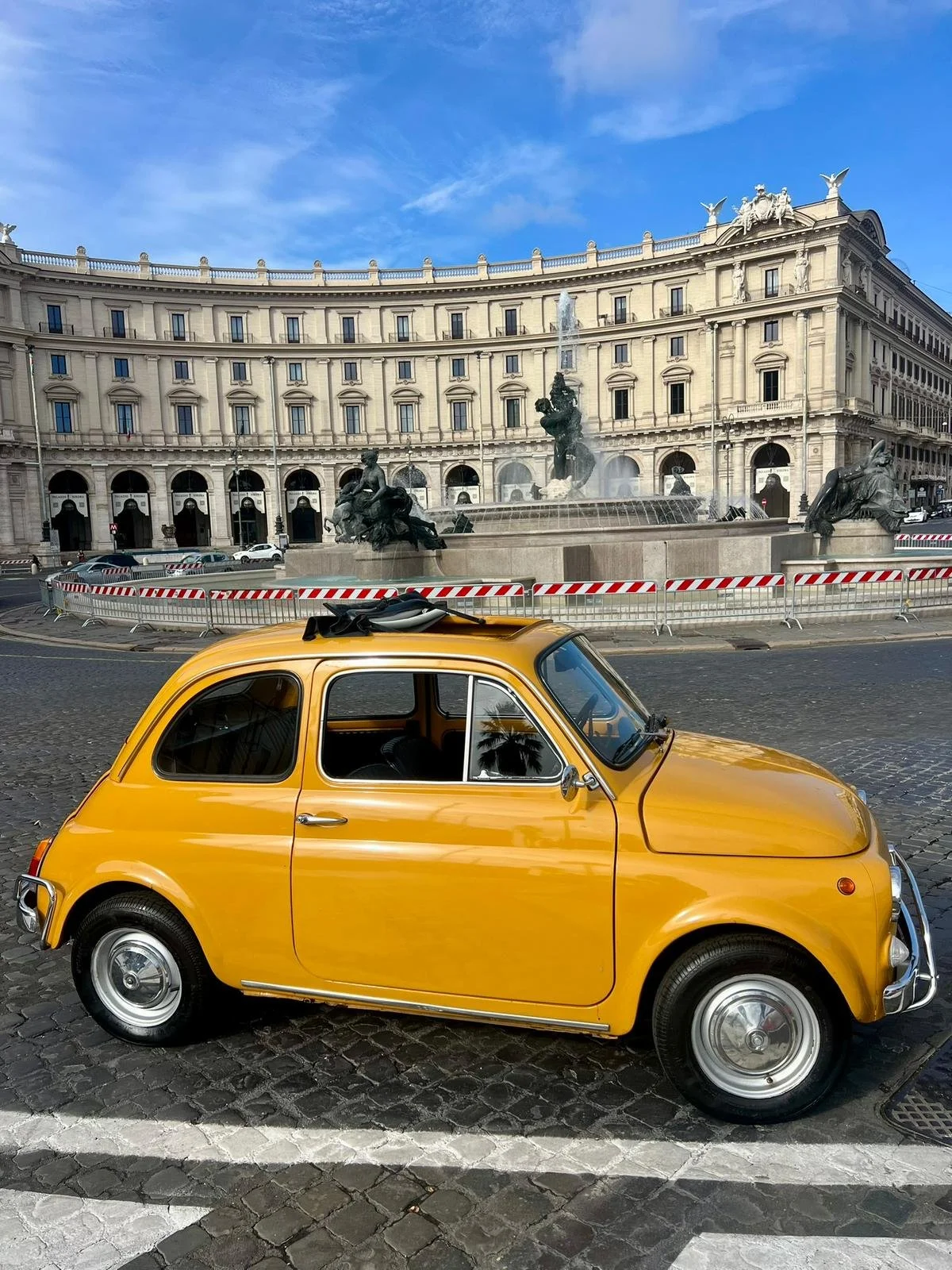 Auto gialla parcheggiata di fronte a una fontana con sculture, in una piazza con un grande edificio storico dietro, sotto un cielo blu.