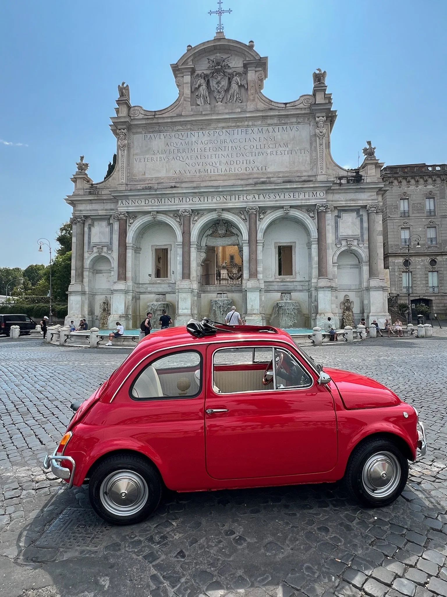 Una piccola auto rossa, tipicamente un'auto d'epoca, parcheggiata su una superficie di pietre davanti a una grande chiesa storica con una facciata decorata e dettagli architettonici elaborati.