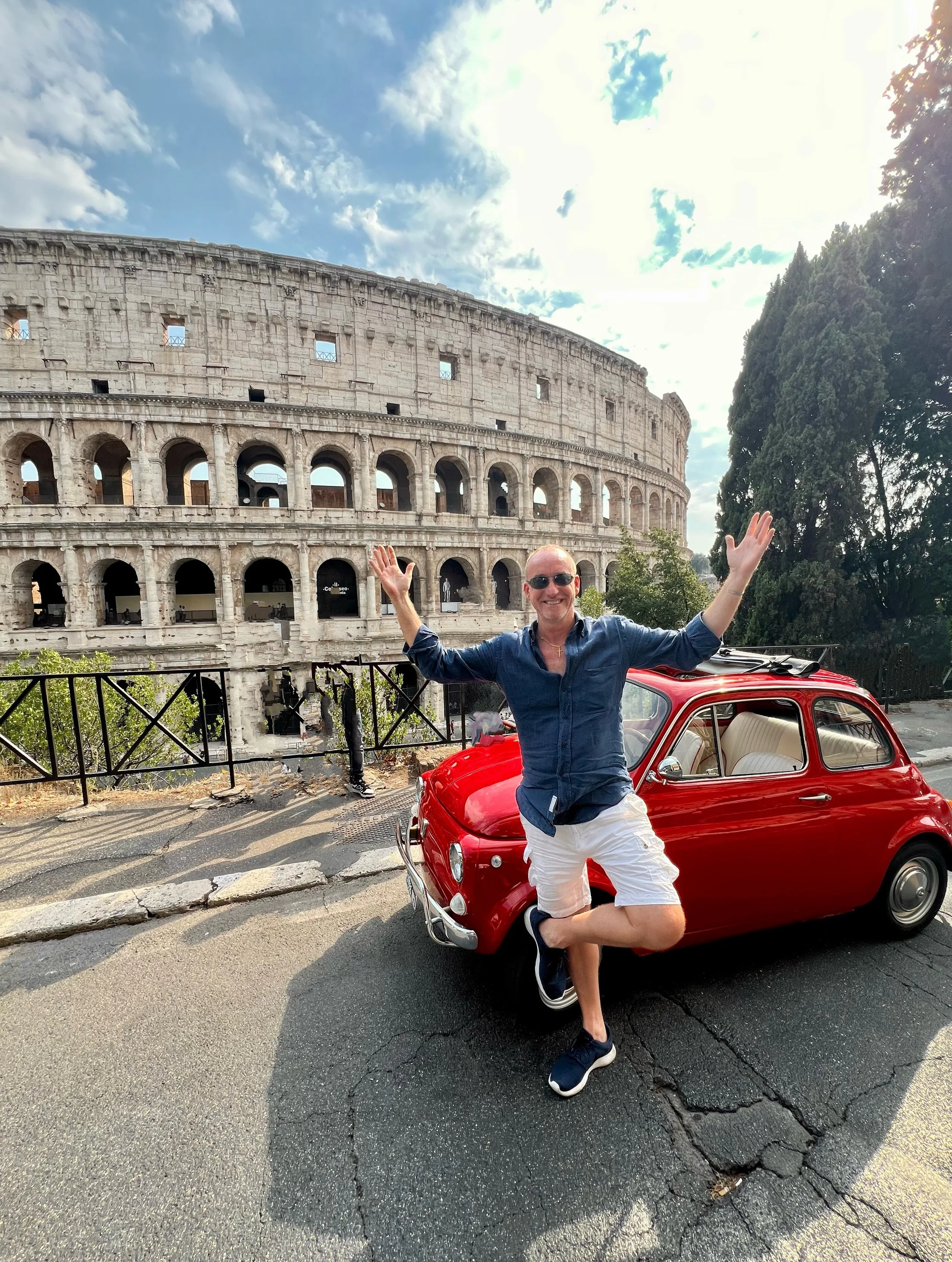 Uomo sorridente con abbigliamento casual e occhiali da sole, con braccia aperte, in piedi accanto a una piccola auto rossa, di fronte al Colosseo a Roma in una giornata soleggiata con alcune nuvole nel cielo.