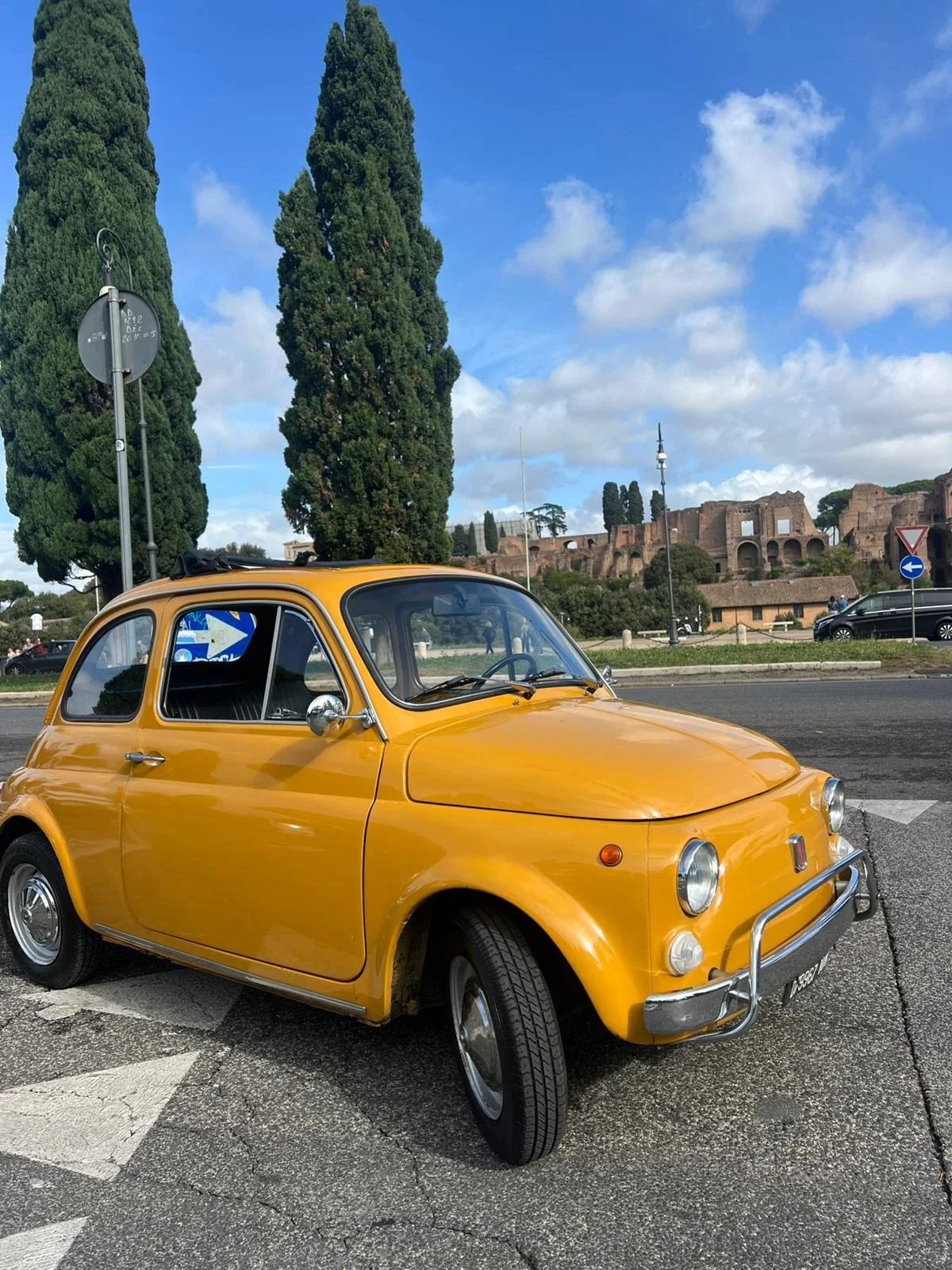 Auto d'epoca di colore giallo parcheggiata su strada con sfondo di alberi e rovine di antica città romana