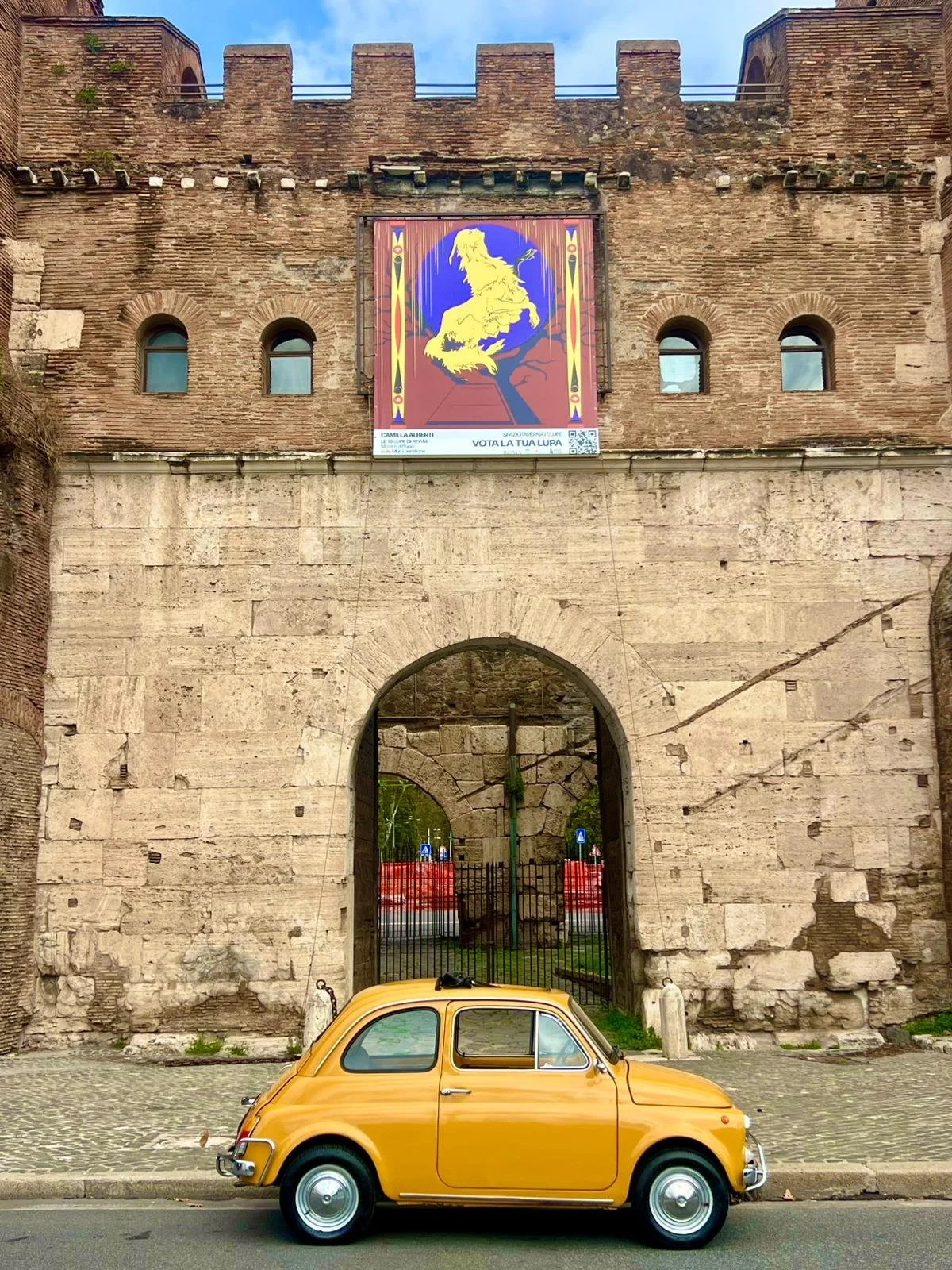 Vettura gialla vintage parcheggiata davanti a un antico muro di pietra con un grande portale. Sopra l'arcata, un grande poster con un leone stilizzato giallo su sfondo blu e rosso.