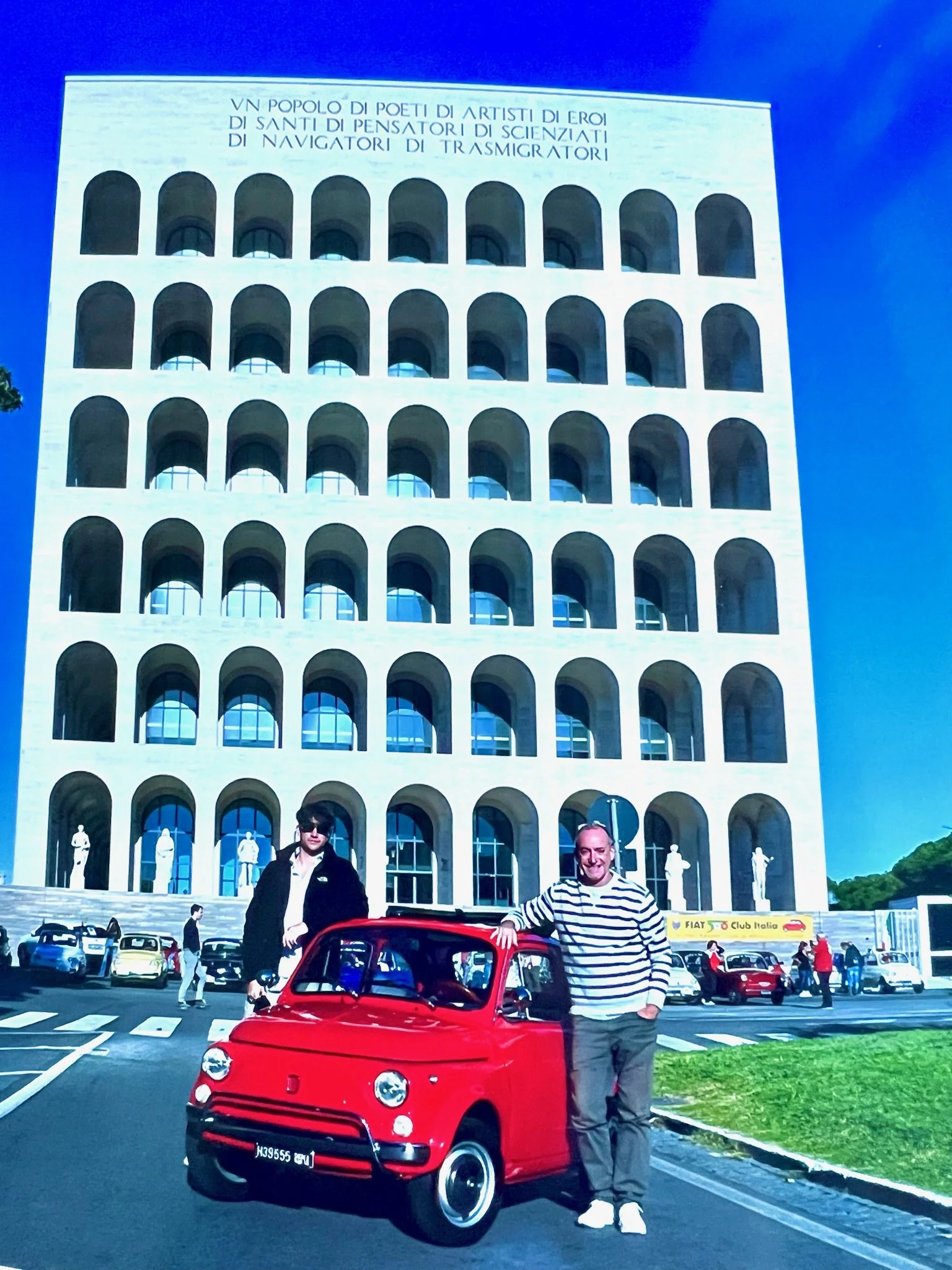 Due uomini davanti a un'auto rossa vintage, con un edificio in stile antico sullo sfondo, in una piazza con altre auto e persone.