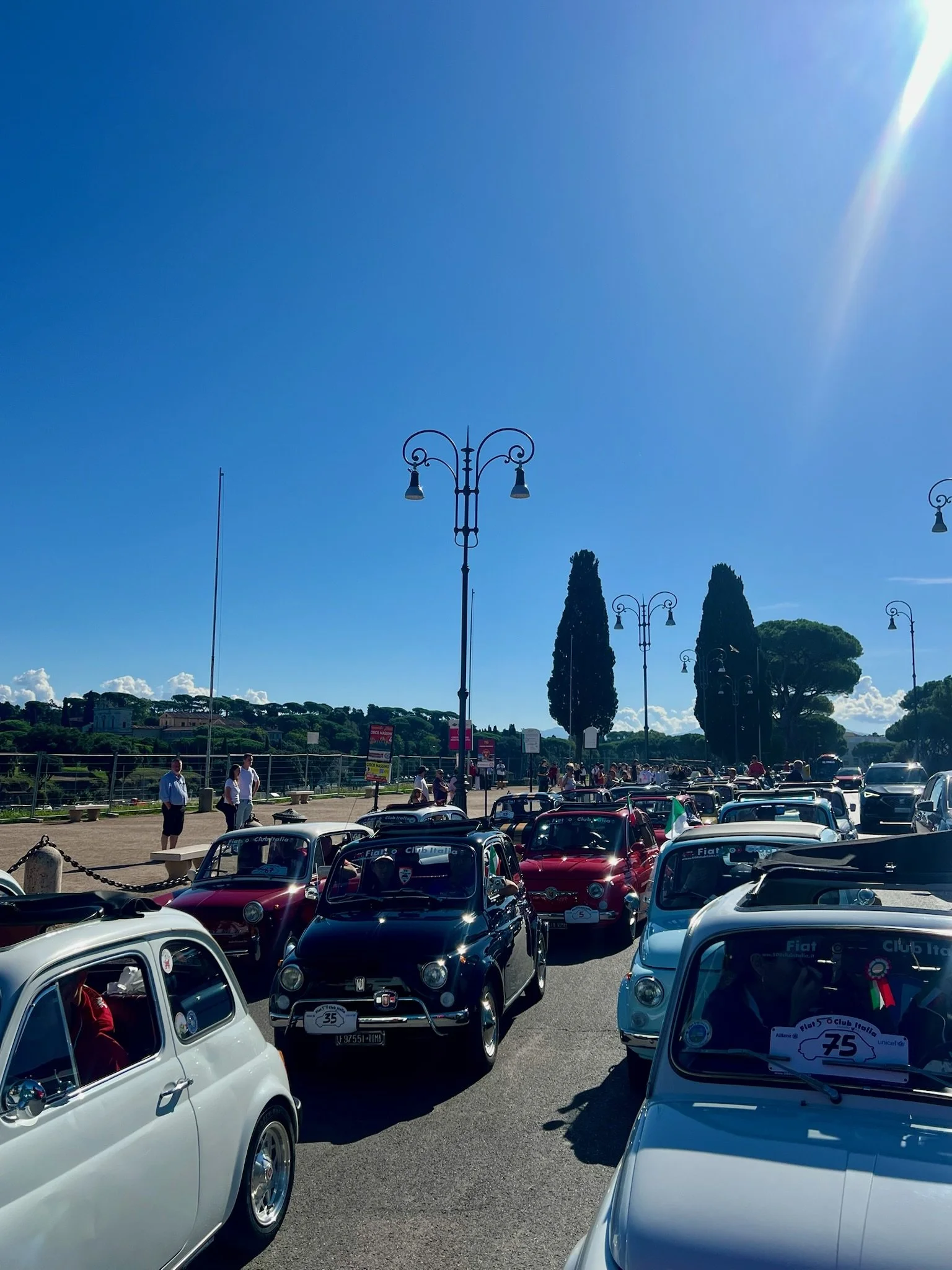 Parco con molte auto d'epoca Fiat, persone e alberi sotto un cielo azzurro soleggiato.