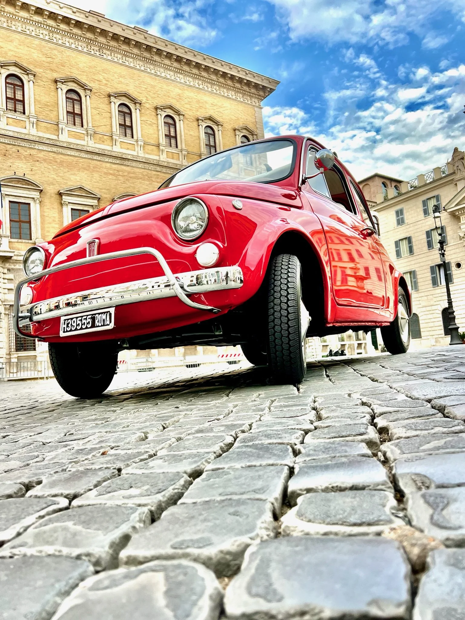 Una macchina d'epoca rossa parcheggiata su una strada di pietra in una piazza storica, con edifici antichi e un cielo azzurro con nuvole.