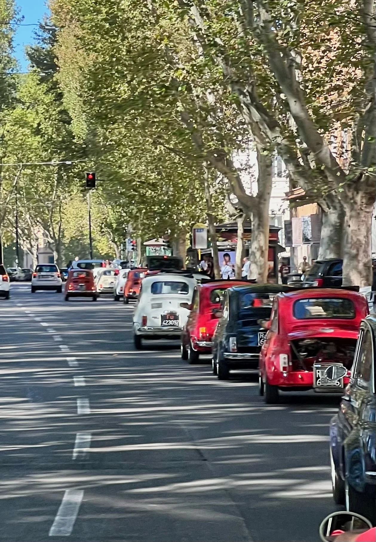 Strada cittadina con auto d'epoca parcheggiate e semaforo rosso, alberi lungo il marciapiede e qualche persona ai lati della strada.