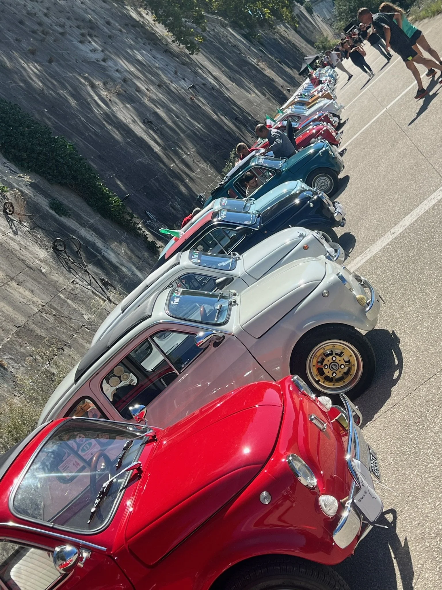 Fila di microcar di vari colori parcheggiate lungo una strada, con alcune persone che le osservano o le scattano fotografie.