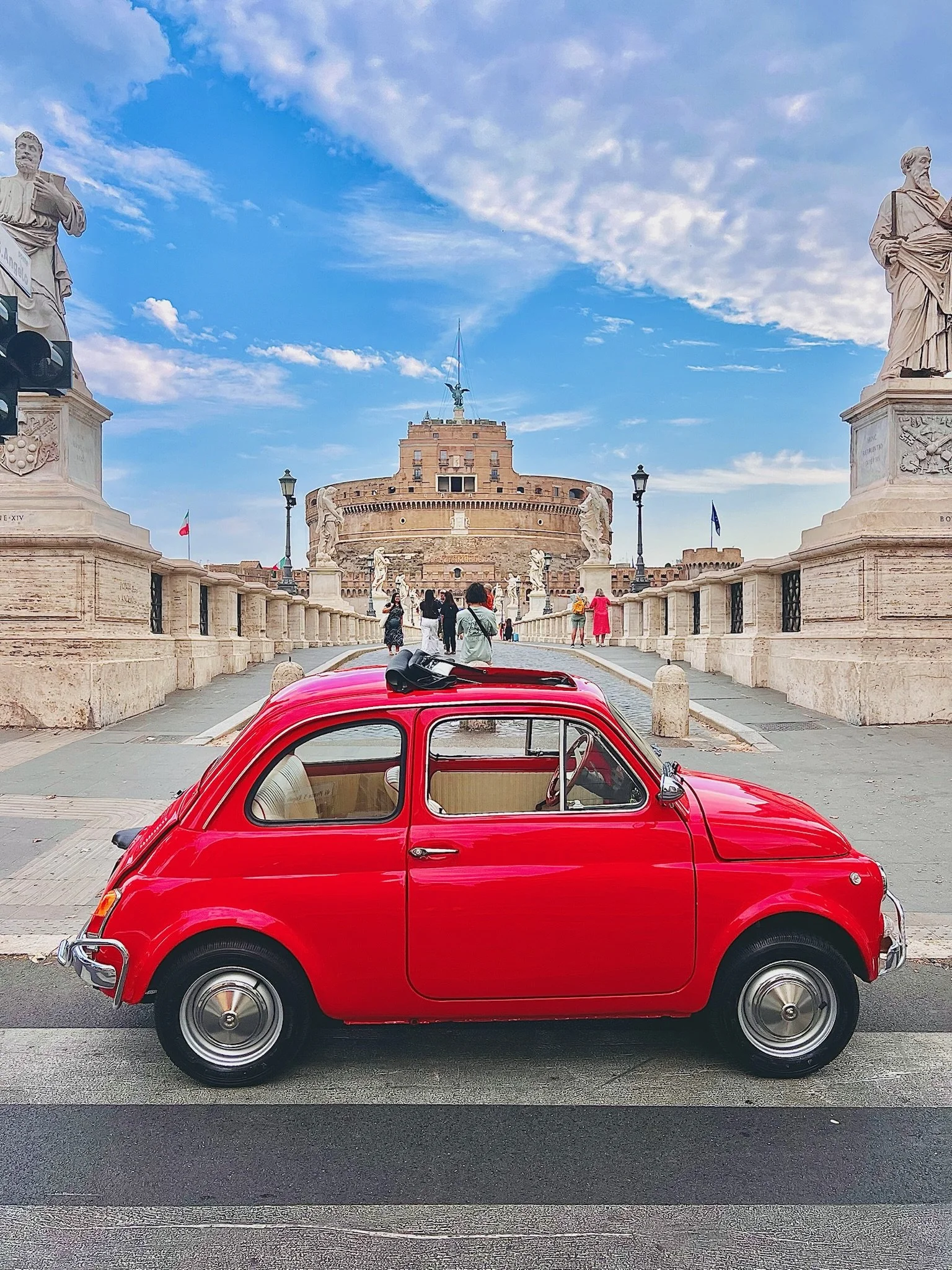 Automobile rossa vintage parcheggiata di fronte a Castel Sant'Angelo a Roma, con il sky e le statue storiche visibili.