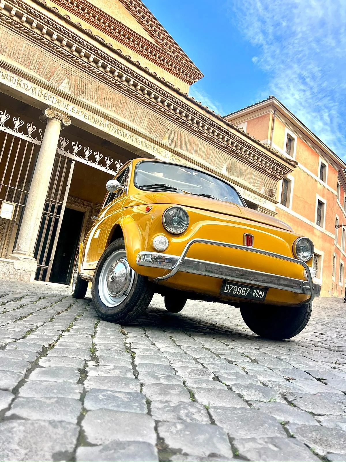 Auto d'epoca gialla parcheggiata su un'originaria strada di ciottoli, davanti a un edificio storico con colonne e altoscritti in latino, sotto un cielo blu con nuvole sparse.