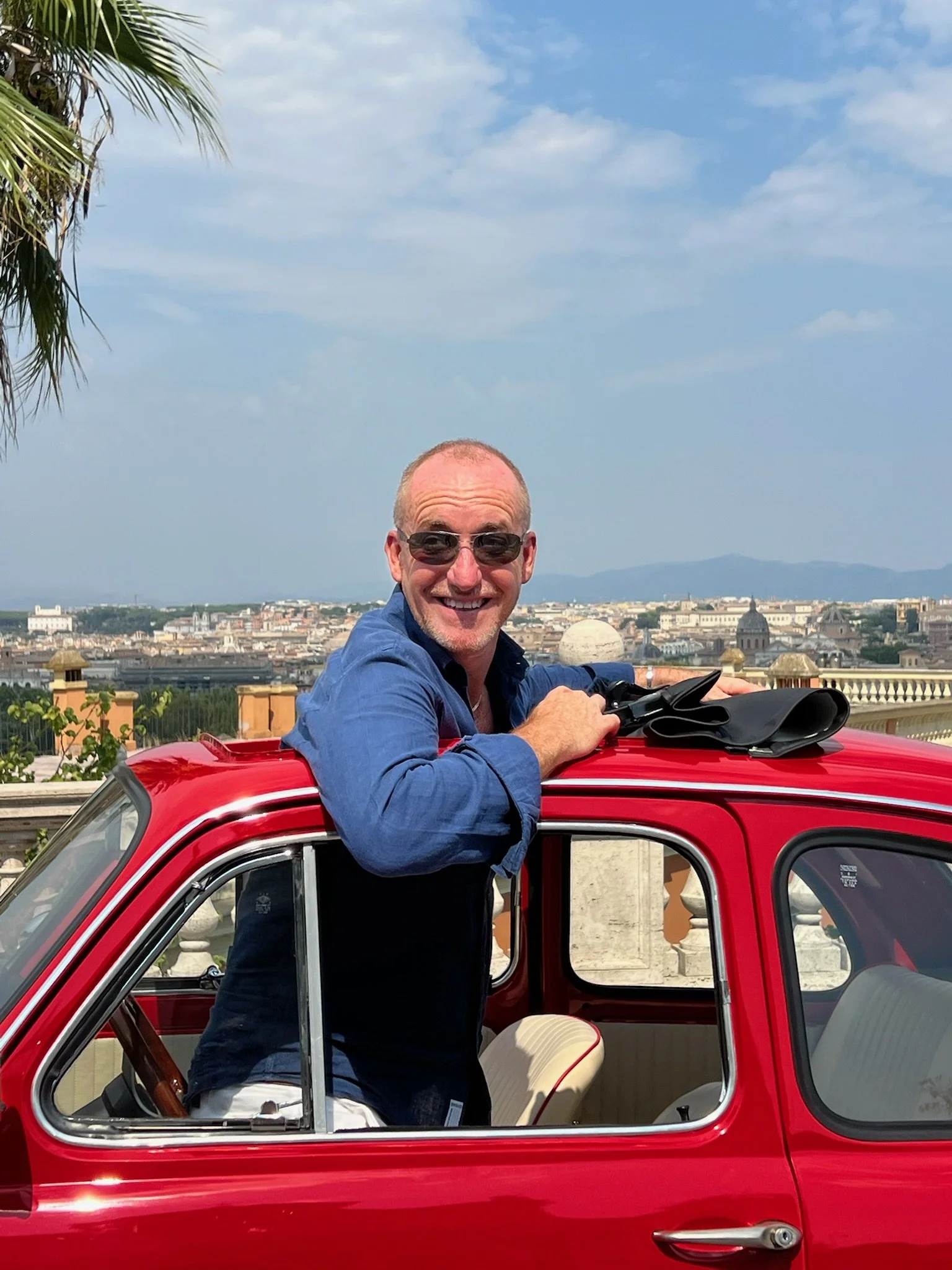 Uomo sorridente con occhiali da sole che sorregge un'auto rossa di piccole dimensioni con un panorama urbano sullo sfondo.
