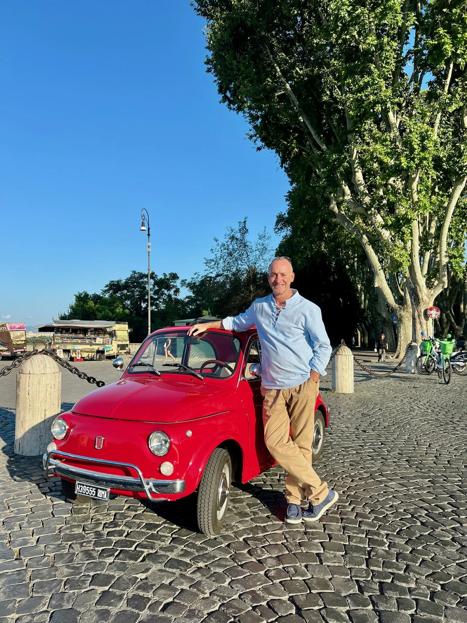 Uomo abbraccia una piccola auto rossa in un'area all'aperto soleggiata circondata da alberi e altri veicoli.