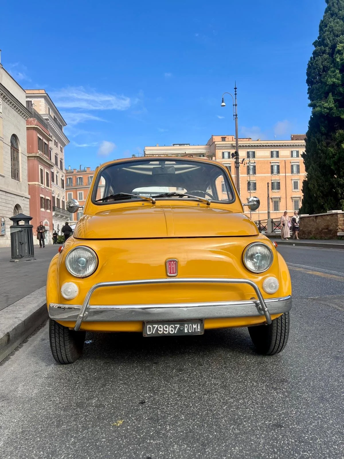 Auto giallo Fiat 500 con targa Roma, parcheggio sulla strada in una zona urbana con edifici storici e persone che passeggiano