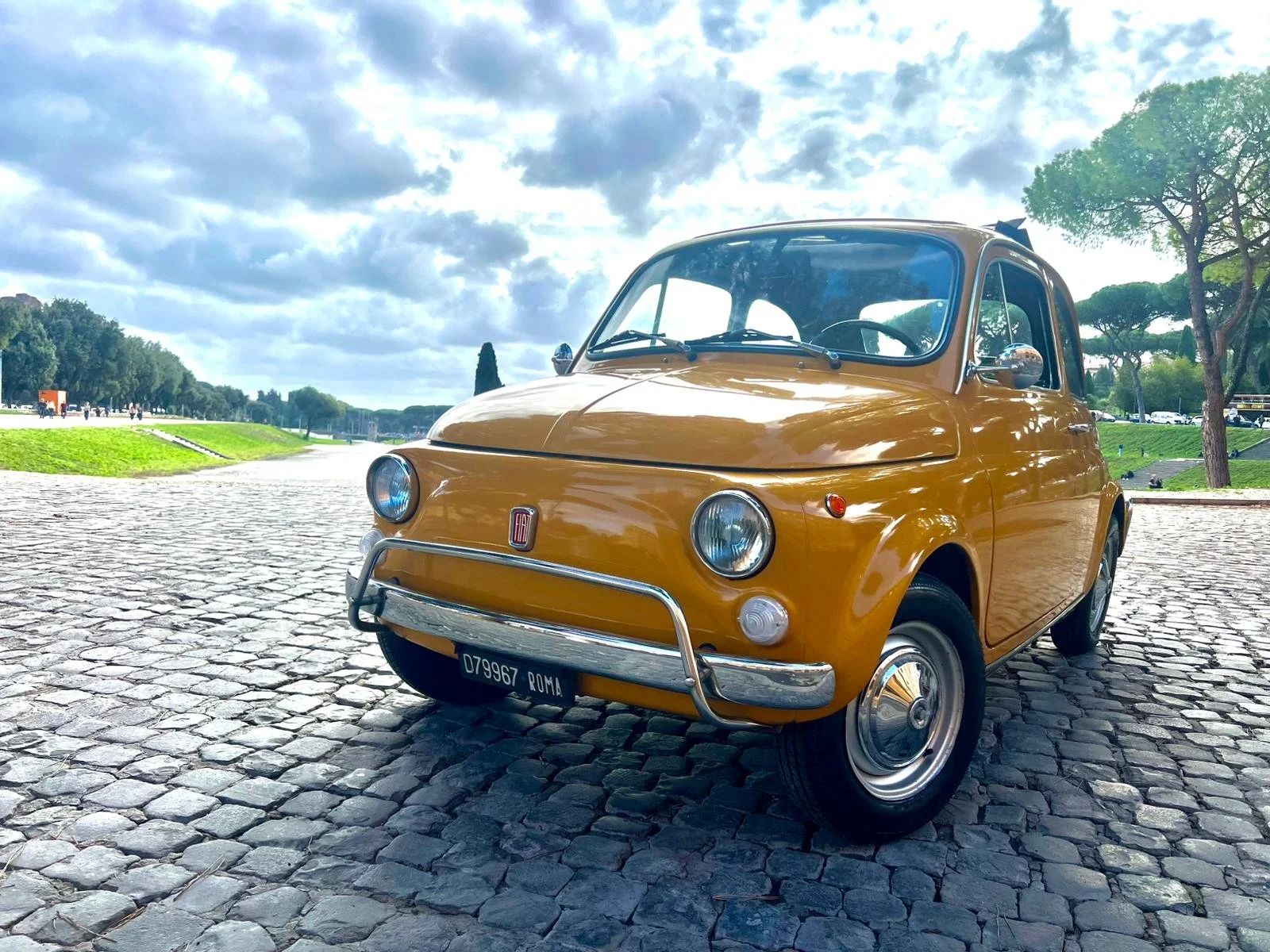 Auto d'epoca Fiat 500 di colore giallo parcheggiato su una strada di ciottoli, con alberi e cielo nuvoloso sullo sfondo.