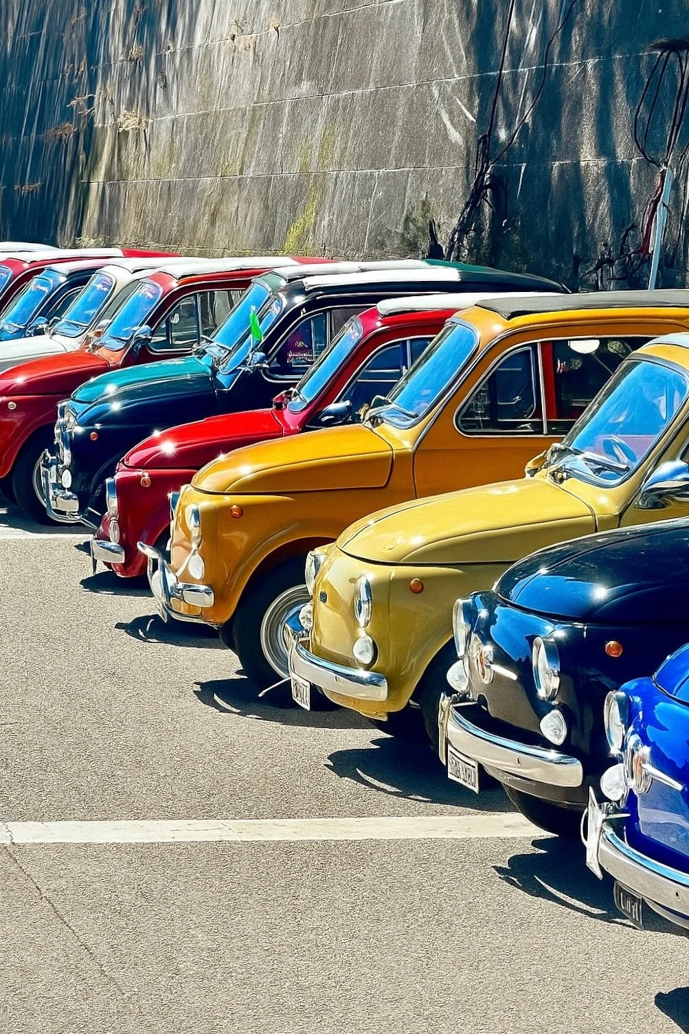 Una fila di auto d'epoca di vari colori, tra cui rosso, nero, giallo, blu e bianco, parcheggiate lungo una strada con un muro di pietra sullo sfondo.